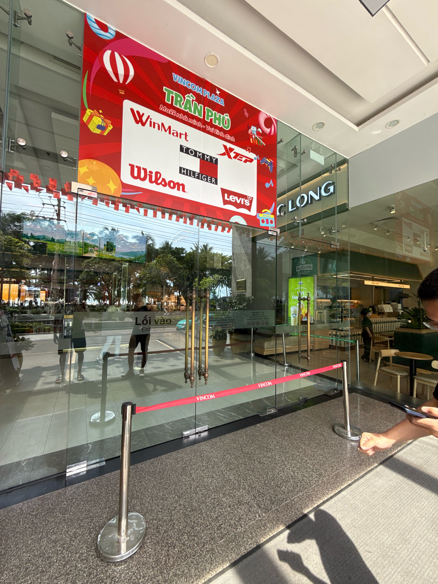 This image depicts the exterior of a Vincom Plaza shopping center in Nha Trang, Vietnam. The foreground features a glass entrance with stanchions and a red velvet rope, suggesting a queuing area. Reflections on the glass show trees, palm trees, and the sky. A large red banner with graphics and text, including "VINCOM PLAZA TRẦN PHÚ" and brand logos such as "WinMart", "TOMMY HILFIGER", and "Levi's", is prominently displayed on the left side of the entrance. To the right of the entrance, the Vincom Plaza logo and "CLONG" are visible on the glass facade. Inside, a store or cafe with tables, chairs, and a counter is visible. A person is partially visible on the right side of the image, holding a smartphone. The overall scene suggests a commercial area with an emphasis on shopping and leisure.