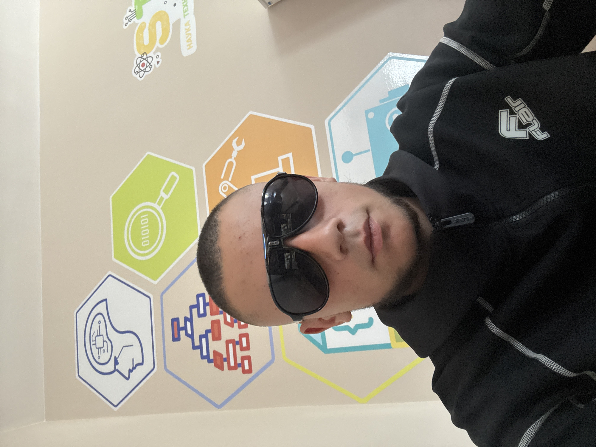 A man with sunglasses and a black jacket is shown in the image. Behind him is a wall decorated with colorful hexagonal icons related to science and technology. Based on the writing on the wall, the location is identified as Elhovo, Bulgaria.