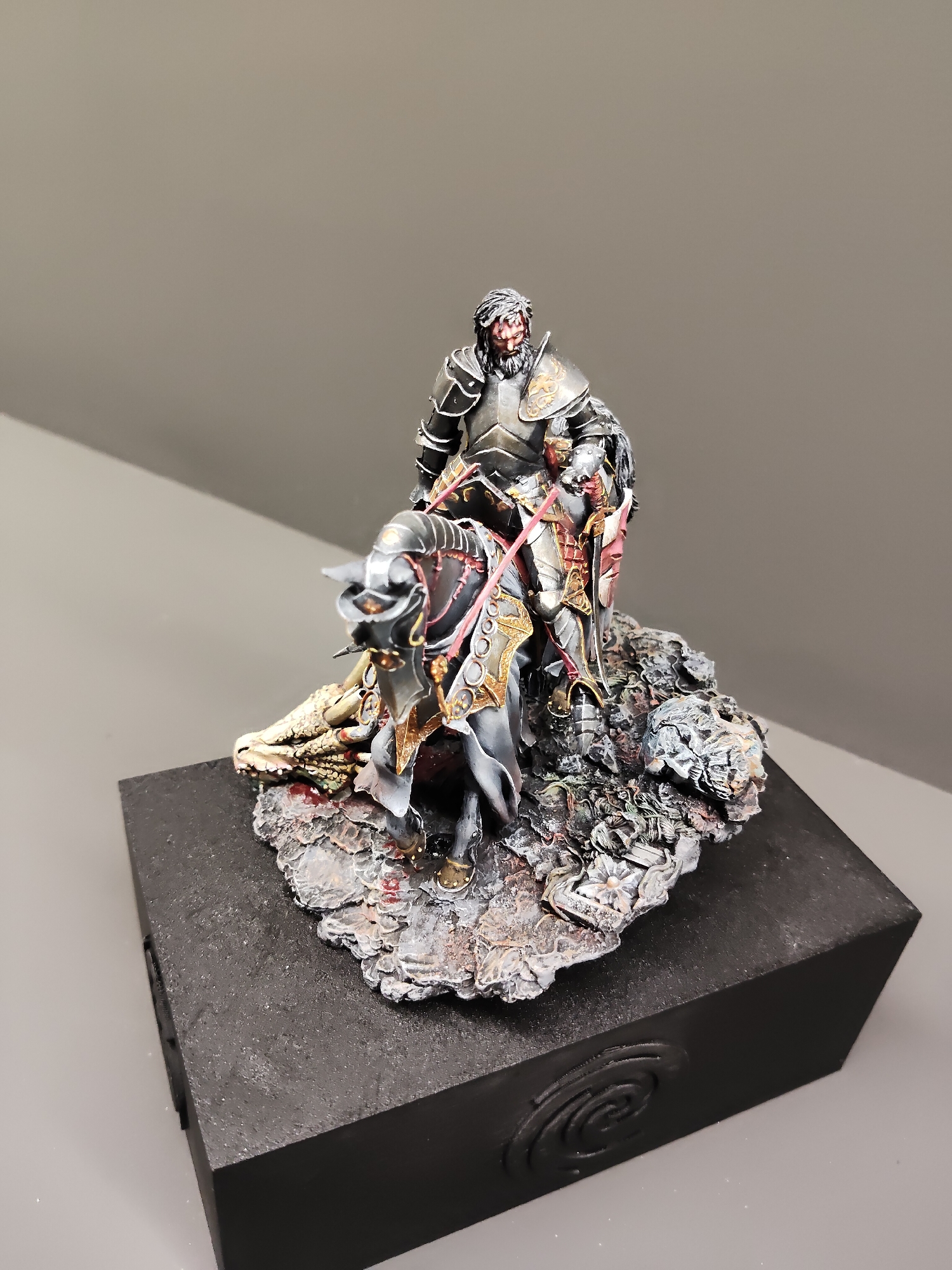 A meticulously crafted miniature diorama depicts a medieval warrior clad in dark, ornate armor, standing over a fallen dragon. The warrior has a beard and a stern expression, with red accents on his armor and a pink sword hilt. The dragon's body is visible, with scales and what appears to be blood spatter on the rocky terrain. The diorama is mounted on a black rectangular base with a embossed circular design. The background is a plain grey wall, and the lighting suggests an indoor setting. There is no indication of the location being Singapore.