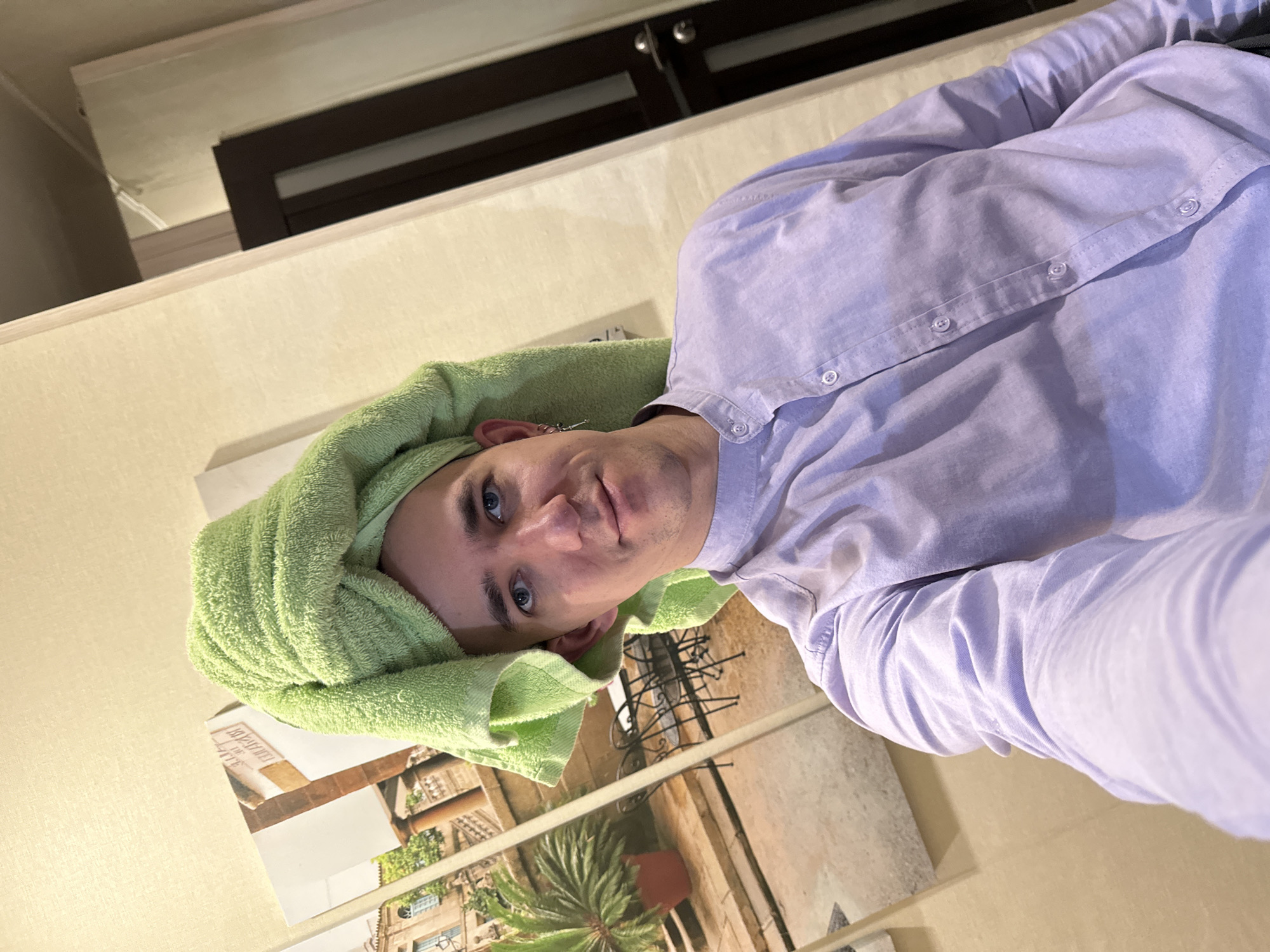 A selfie of a man indoors, wearing a light purple, button-down shirt and a green towel wrapped around his head. He is looking at the camera. Behind him, there is a wall with a decorative picture showing buildings and plants. A dark brown cabinet or wardrobe is visible in the upper background. The location is likely a domestic setting, possibly in Saratov, Russia.