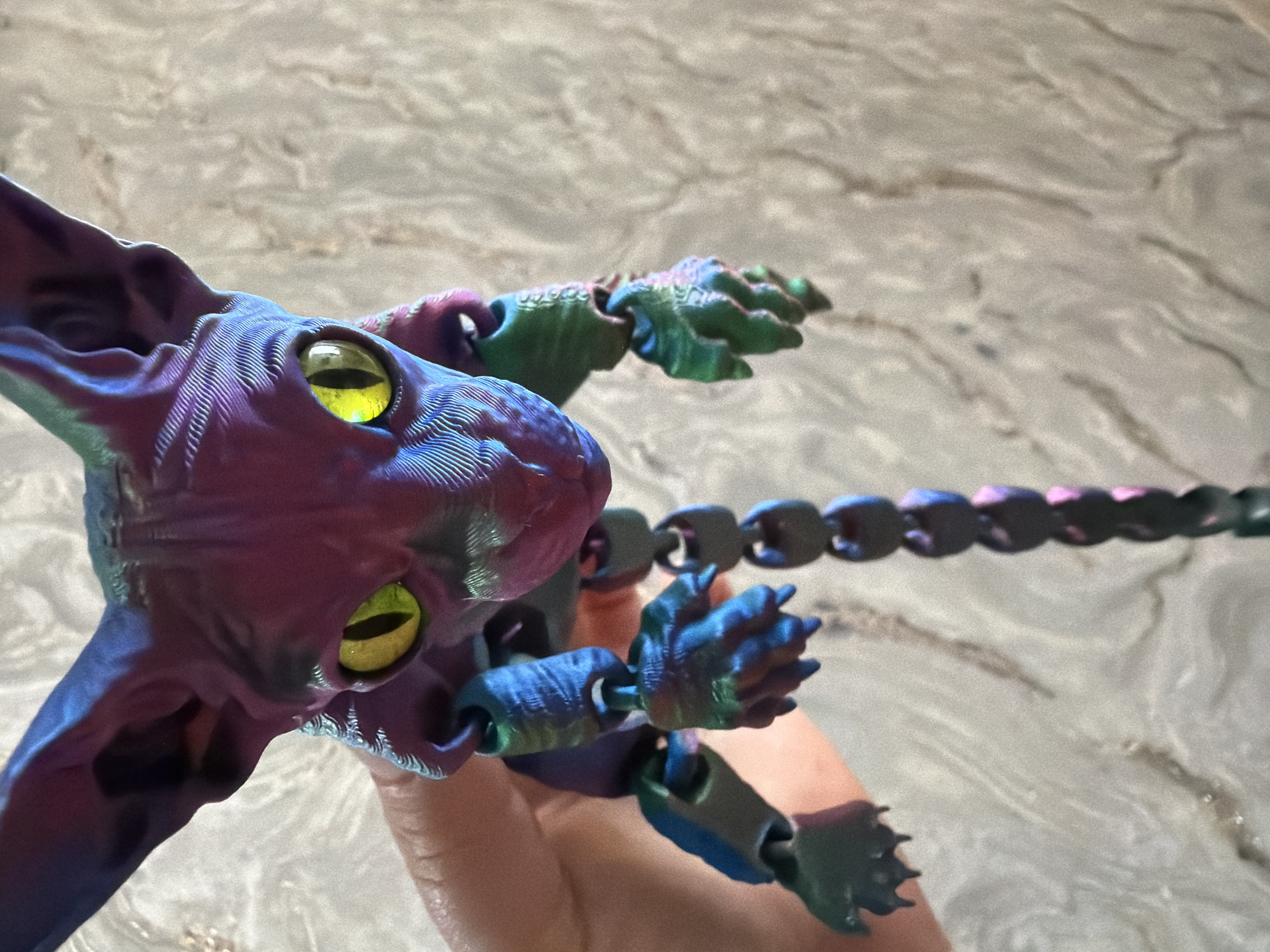 An articulated 3D printed toy resembling a Sphynx cat is held in a person's left hand against a textured, light-colored background. The toy has large, yellow-green eyes with black pupils, and its body features a gradient color transition from purple to blue and green. The cat's tail is a segmented chain, also displaying the same color gradient. The person's hand is partially visible, supporting the toy. The background appears to be a wall with an abstract pattern. The scene is brightly lit. The location is unknown.