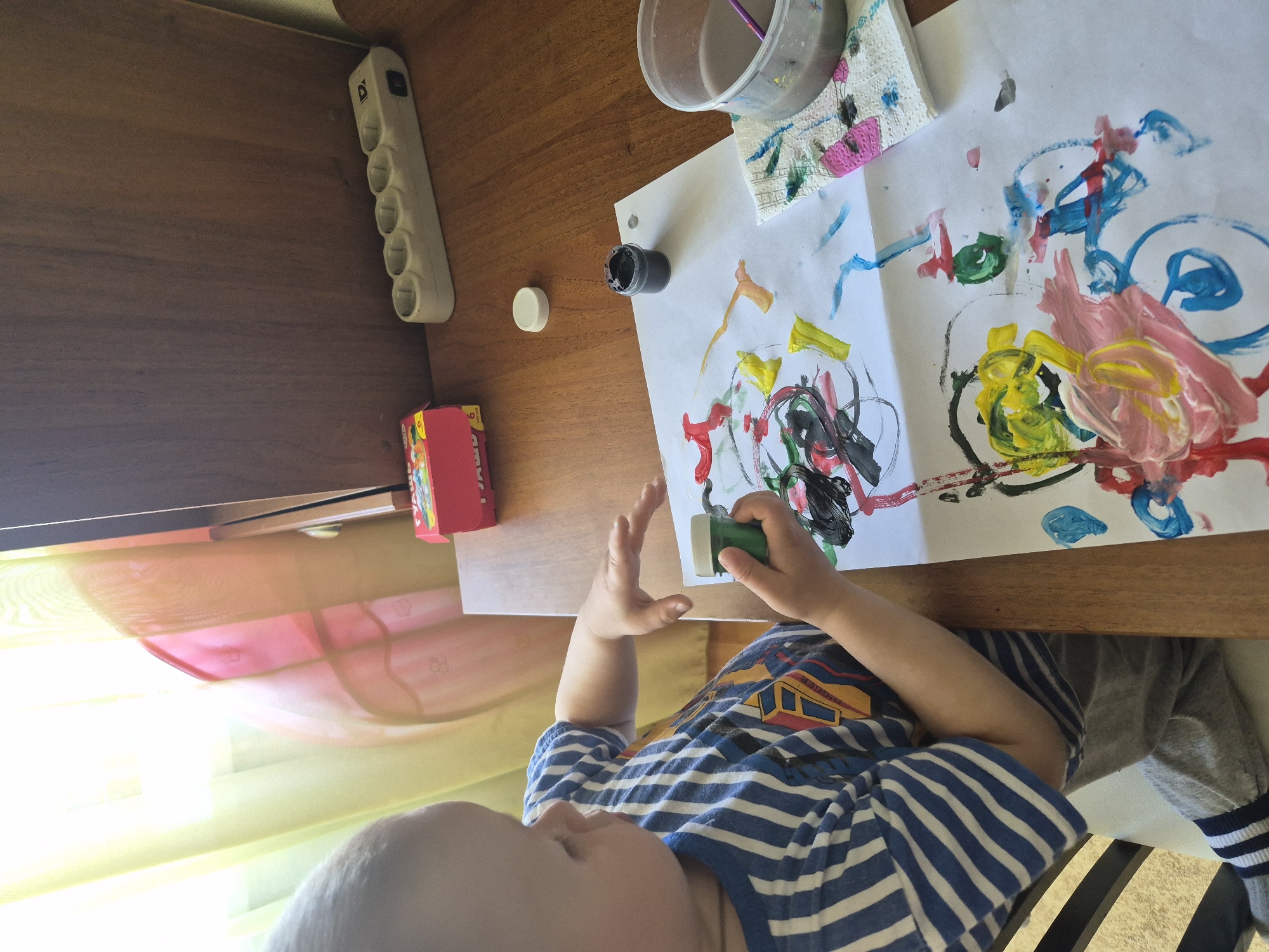 A child sits at a wooden table and paints on a white piece of paper. The child is wearing a blue and white striped shirt with a cartoon train on it and gray pants. The paper contains colorful paint splatters and crude drawings. On the table, there is a container of water, a paint brush, a small jar of black paint, and a red box. An electrical outlet strip is mounted on the edge of the table. The image was taken from directly above the child and table. The location is a home, likely in Tobolsk, Russia. The time is daytime due to sunlight entering in the bottom left portion of the frame.