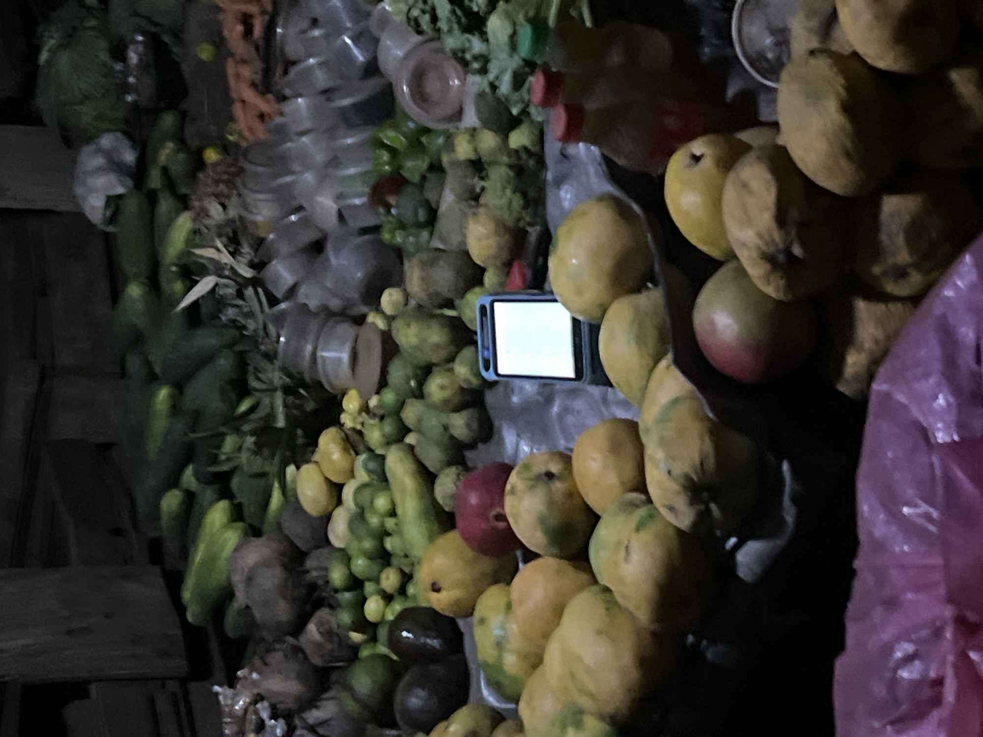 The image shows a dark, possibly indoor, market scene in Kubwa, Nigeria. Various produce items are displayed, including cucumbers, avocados, limes, mangoes, and leafy green vegetables. Discarded plastic containers are visible in the background. A smartphone with a bright screen is placed among the fruits. A purple plastic bag is also visible. The overall lighting is poor, obscuring some details.