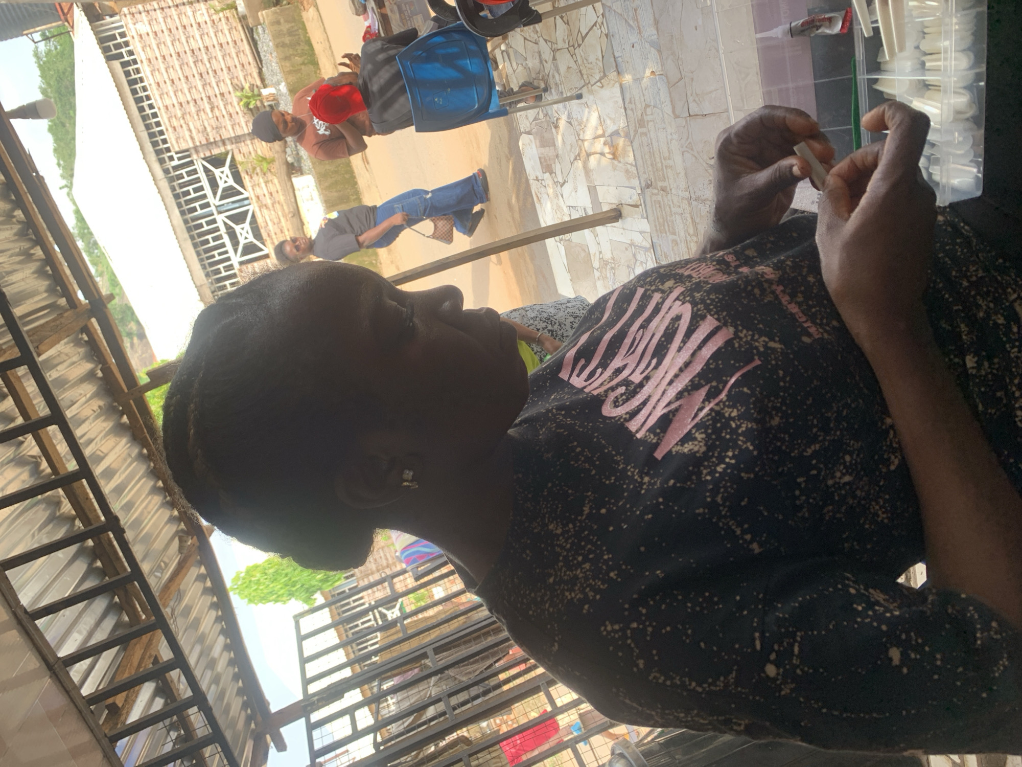 A woman is shown from the chest up, holding a false fingernail in front of a clear plastic container of nails. The woman wears a black t-shirt with a pink design. Outside, blurred people and a building are visible. The location appears to be Abuja, Nigeria.