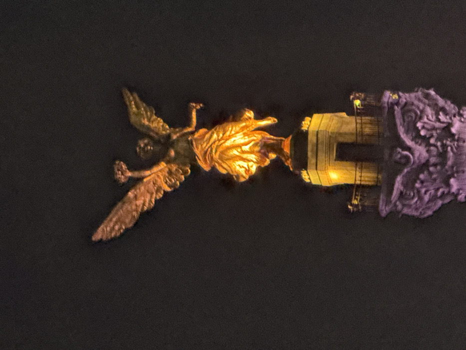 A nighttime view of the Ángel de la Independencia statue in Mexico City. The golden angel figure is depicted in flight, atop a column illuminated in yellow. The base of the monument features detailed carvings with a purple hue, contrasting with the dark background. The entire image is rotated ninety degrees clockwise.
