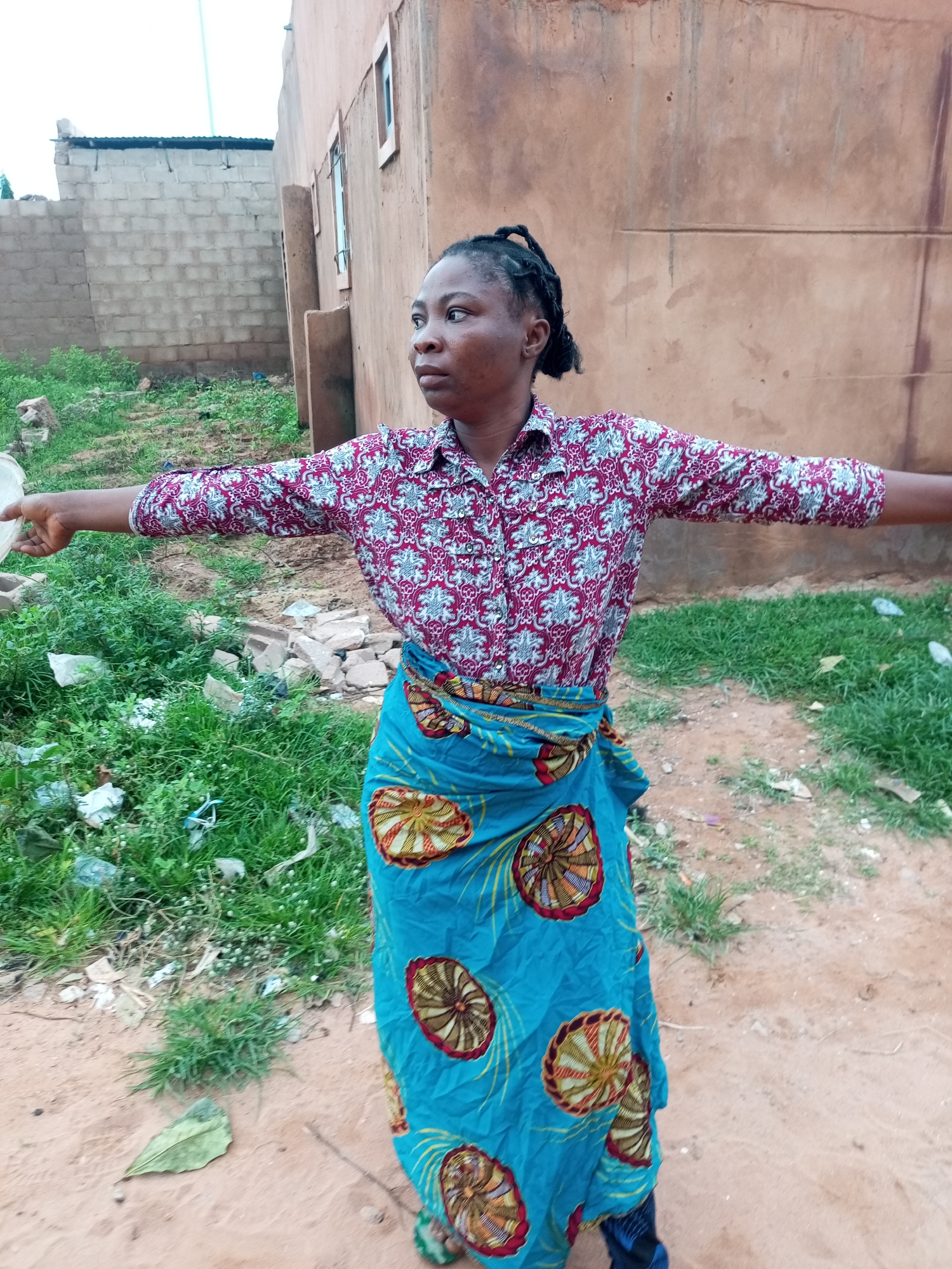 A young woman with braided hair stands with her arms outstretched to the sides, facing slightly to her right. She wears a maroon and white patterned button-down shirt and a bright blue wrap skirt with a repeating circular pattern in yellow, red, and brown. The background consists of a dirt path, overgrown green grass with scattered debris, and two buildings. To the left, a light-colored brick building with a dark roof is visible. To the right, a large, muted orange building with horizontal lines and rectangular window frames dominates the scene. The sky is overcast. The woman appears to be in a semi-urban or rural area, possibly on the outskirts of Tempure, Nigeria, indicated by the style of construction and vegetation.