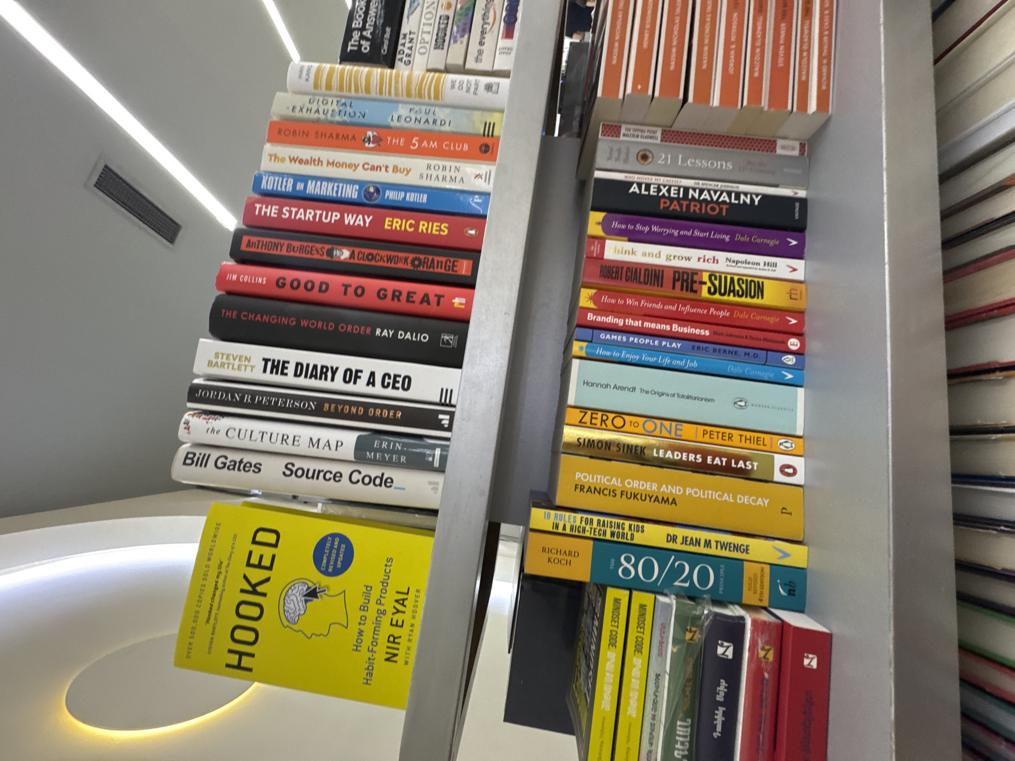 The image shows a close-up of two bookshelves filled with books. The left bookshelf contains titles such as "Hooked", "Source Code", "The Culture Map", "The Diary of a CEO", "Good to Great", "The Startup Way", "The 5 AM Club", and "Digital Exhaustion". The right bookshelf features titles like "21 Lessons", "Alexei Navalny Patriot", "Think and Grow Rich", "Pre-Suasion", "Games People Play", "Zero to One", "Leaders Eat Last", "Political Order and Political Decay", "80/20", and "Mindset Code". The bookshelves appear to be inside a building. There is no information to indicate that the location is Yerevan, Armenia.