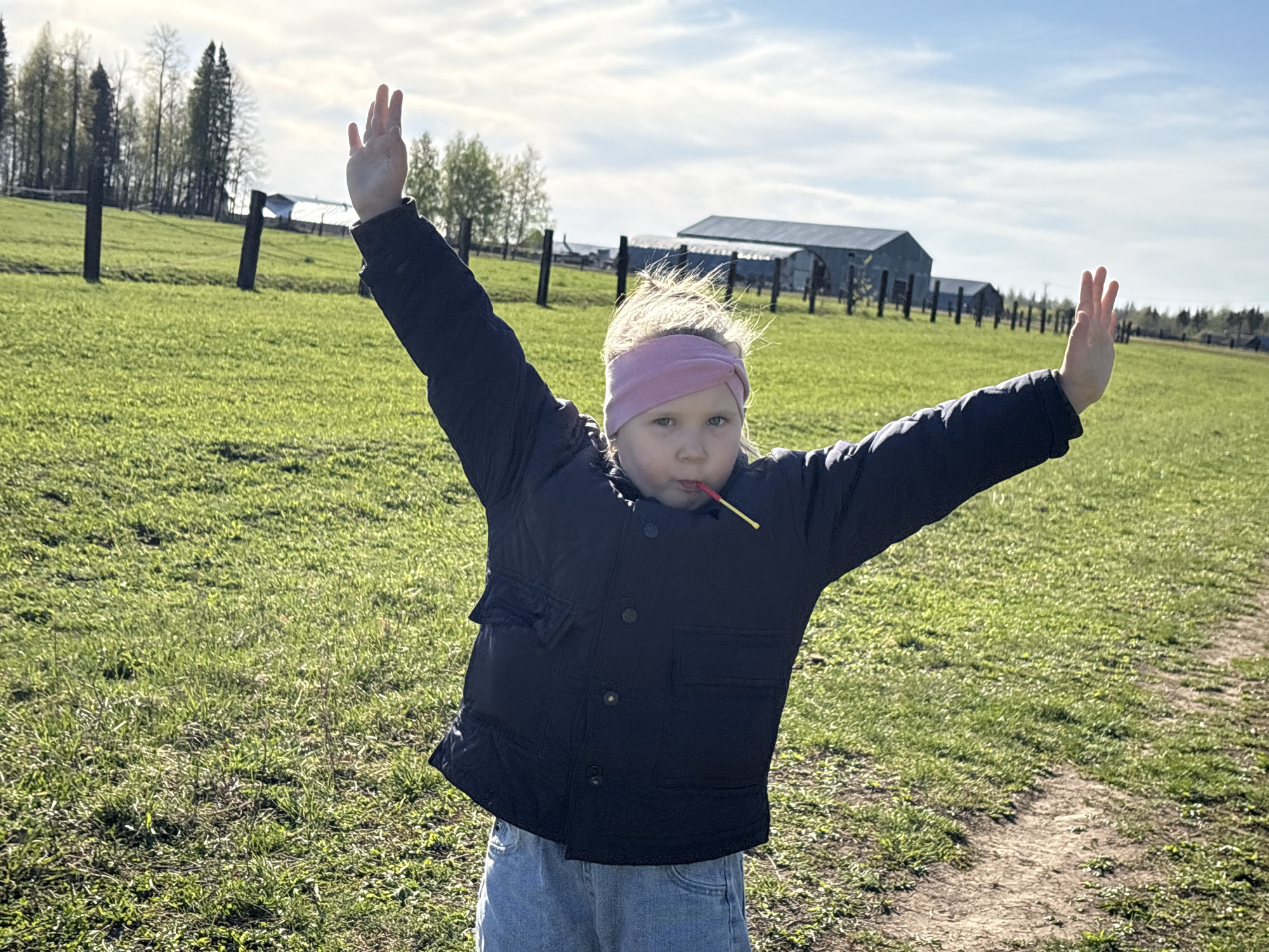 A young girl stands in a grassy field, arms raised above her head, holding a lollipop in her mouth. She wears a dark-colored jacket, blue jeans, and a pink headband. Behind her is a rural scene with a wooden fence, trees, and a large metal building. The sky is bright and cloudy. The photograph was taken outdoors in Азинский, Russia.
