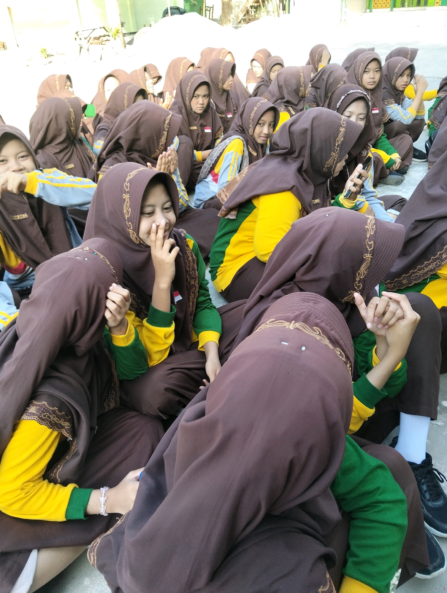 In Dagan, Indonesia, a group of young people wearing brown hijabs and yellow and green long-sleeved shirts are sitting on a paved surface outdoors. The individuals are clustered together, some facing the camera and others with their backs turned. One person in the foreground is holding a dark-colored smartphone. Another person is seen with their hands clasped together. The attire suggests a school uniform. The environment appears to be an outdoor area with a building visible in the background.