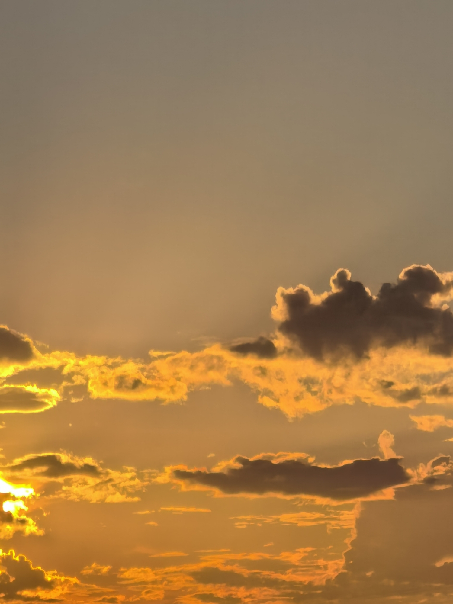 A photograph captures a sunset over a hazy, undefined landscape. The sky is dominated by warm hues of orange and yellow, with scattered clouds catching the last rays of sunlight. Some clouds are a deep, shadowy gray, contrasting with the bright, illuminated portions. The overall impression is one of a serene, end-of-day scene, with the light creating a dramatic and atmospheric effect. The location is unknown.