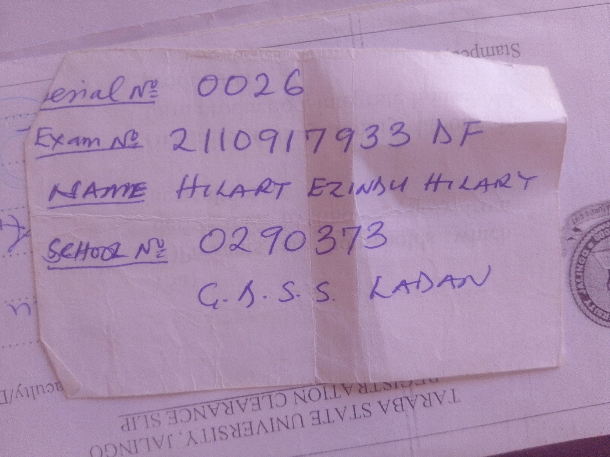 This is a close-up, top-down photograph of a handwritten note placed over a printed document. The note, written in purple ink, contains personal information such as a serial number, exam number, name, and school number. The printed document beneath appears to be an official slip, with visible text like "TARABA STATE UNIVERSITY, JALINGU" and "REGISTRATION CLEARANCE SLIP". There are also stamps present on the printed document, one with "LINGO" and what looks like a year (possibly 800, though it's unclear). The lighting is uneven, with some parts of the note being brighter than others, suggesting the photo was taken indoors with ambient light. The focus is on the handwritten note, with the printed document serving as a background. The style of handwriting suggests a casual, personal context for the note. The overall impression is that of a document related to academic or administrative processes in Nigeria.