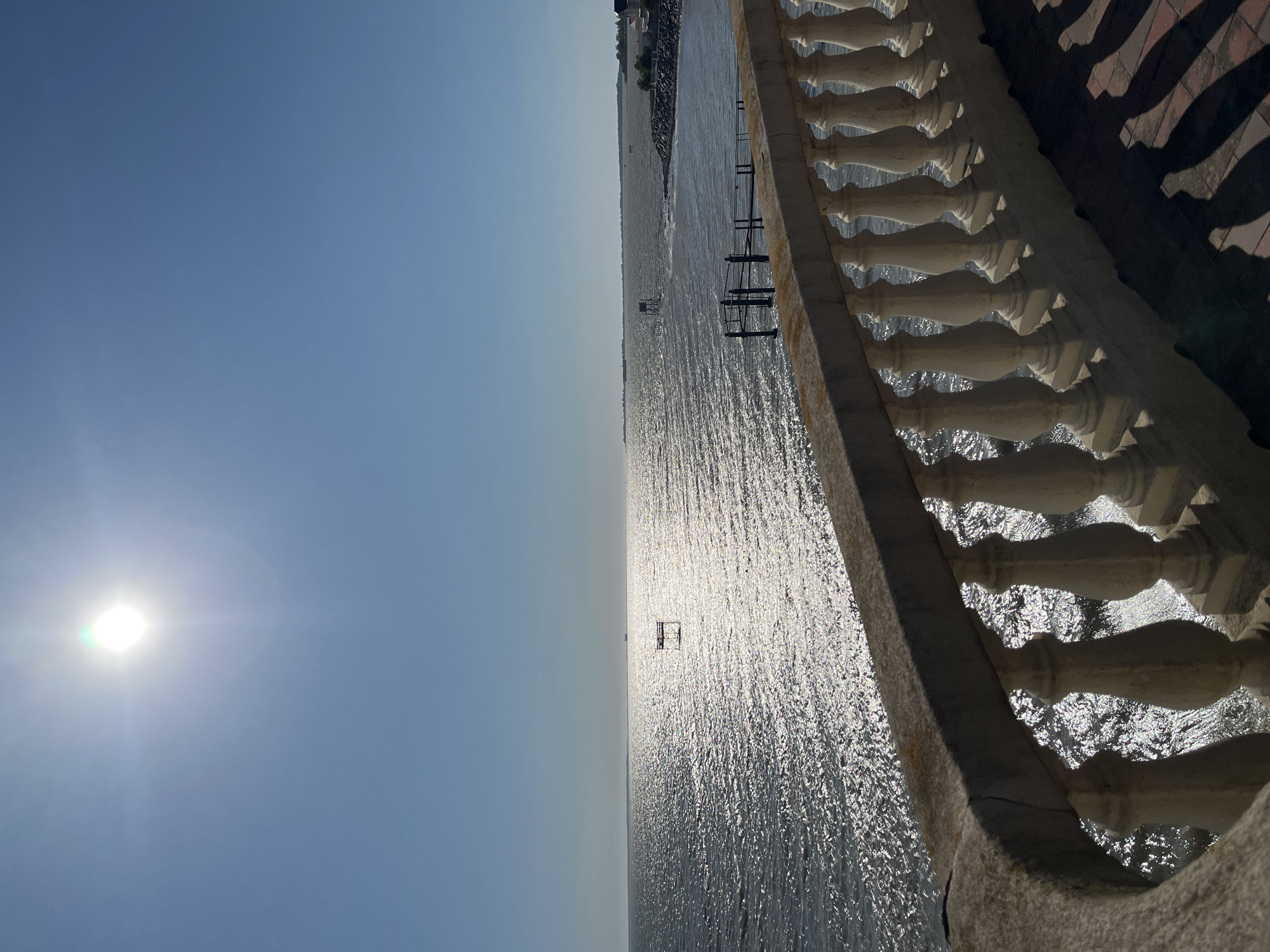 The image depicts a sunlit ocean view from a balcony in Fouras, France. In the foreground, a decorative stone balustrade with white balusters runs along the edge of a tiled patio. The balustrade curves to the left, creating a sense of depth. Beyond the balustrade, the vast ocean stretches to the horizon, its surface shimmering under the bright sunlight. Two small wooden structures on stilts are visible in the water, along with a pier extending from the shore on the right. The sky is a clear, pale blue. The overall scene conveys a peaceful, coastal atmosphere.