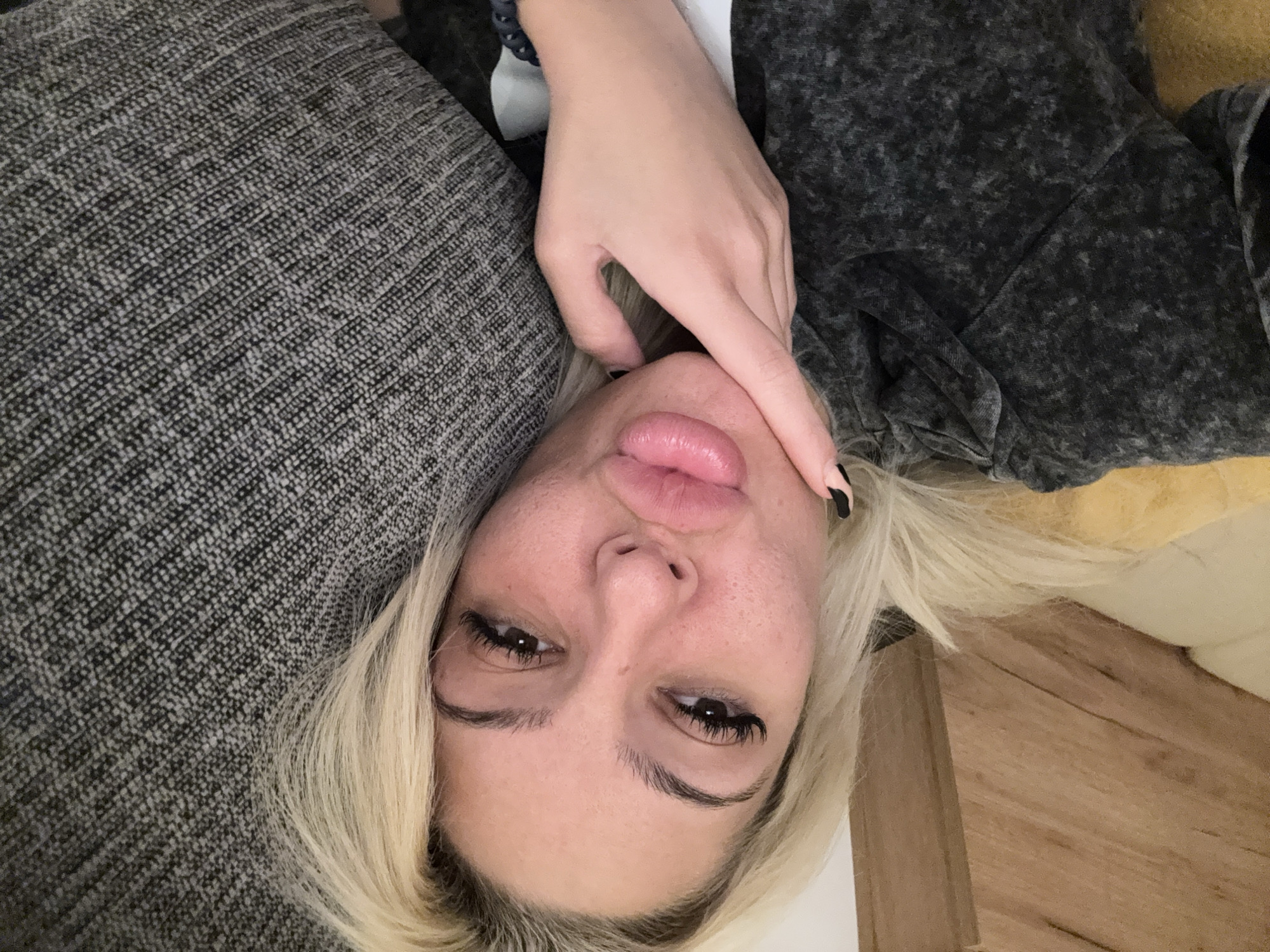 The image is a rotated, close-up portrait of a young woman lying down in an indoor setting. Her blonde hair frames her face, which is slightly tilted. She has dark brown eyes with prominent eyelashes and defined eyebrows. Her full lips are puckered in a "kissy face" or pout. One hand is positioned near her chin and mouth, with black nail polish visible on her fingernails.
She is dressed in a dark gray, mottled or acid-wash textured garment, possibly a casual top like a sweatshirt or hoodie. To her side, a coarse, woven gray fabric, likely a cushion or sofa upholstery, is visible. Adjacent to her head and hair, a lighter, creamy-yellow fabric and a wooden surface (possibly furniture or flooring) are present. A bracelet can be seen on her wrist.
The lighting suggests an indoor environment. Her expression appears casual and playful. There is no visible text. The setting is likely a relaxed living space in Sofia, Bulgaria.