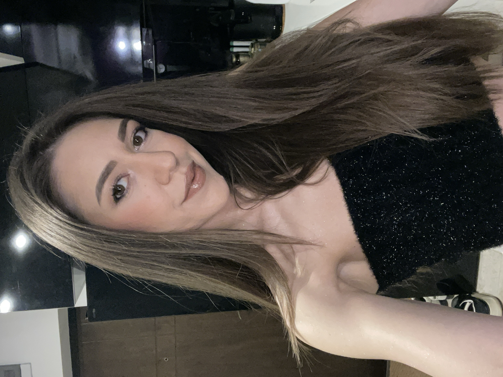 A woman is posing in a vertical portrait shot. She has light brown hair, hazel eyes, and fair skin. She is wearing a black, sparkly top. The background includes dark cabinets or furniture, possibly in a kitchen or bathroom setting. No information on the location in Sofia, Bulgaria, is possible from this image alone.