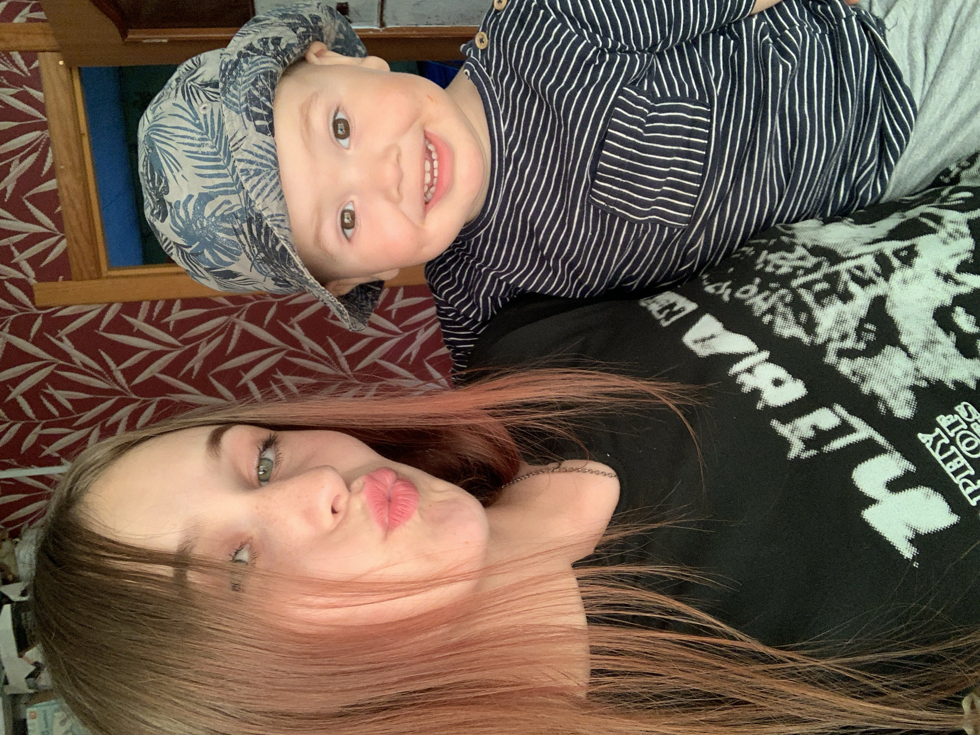 A woman with reddish-brown hair lies on a bed with a patterned, dark-colored sheet. She has fair skin, green eyes, and is making a kissing face toward the camera. She wears a black t-shirt with light-colored lettering. A young child wearing a patterned cap and a navy-and-white striped shirt lies on the woman's shoulder. The child is smiling and looking at the camera. A wooden frame is visible in the background, next to a patterned wall with reddish-brown and white colors. The location is not specified as Фоки, Russia in the image.