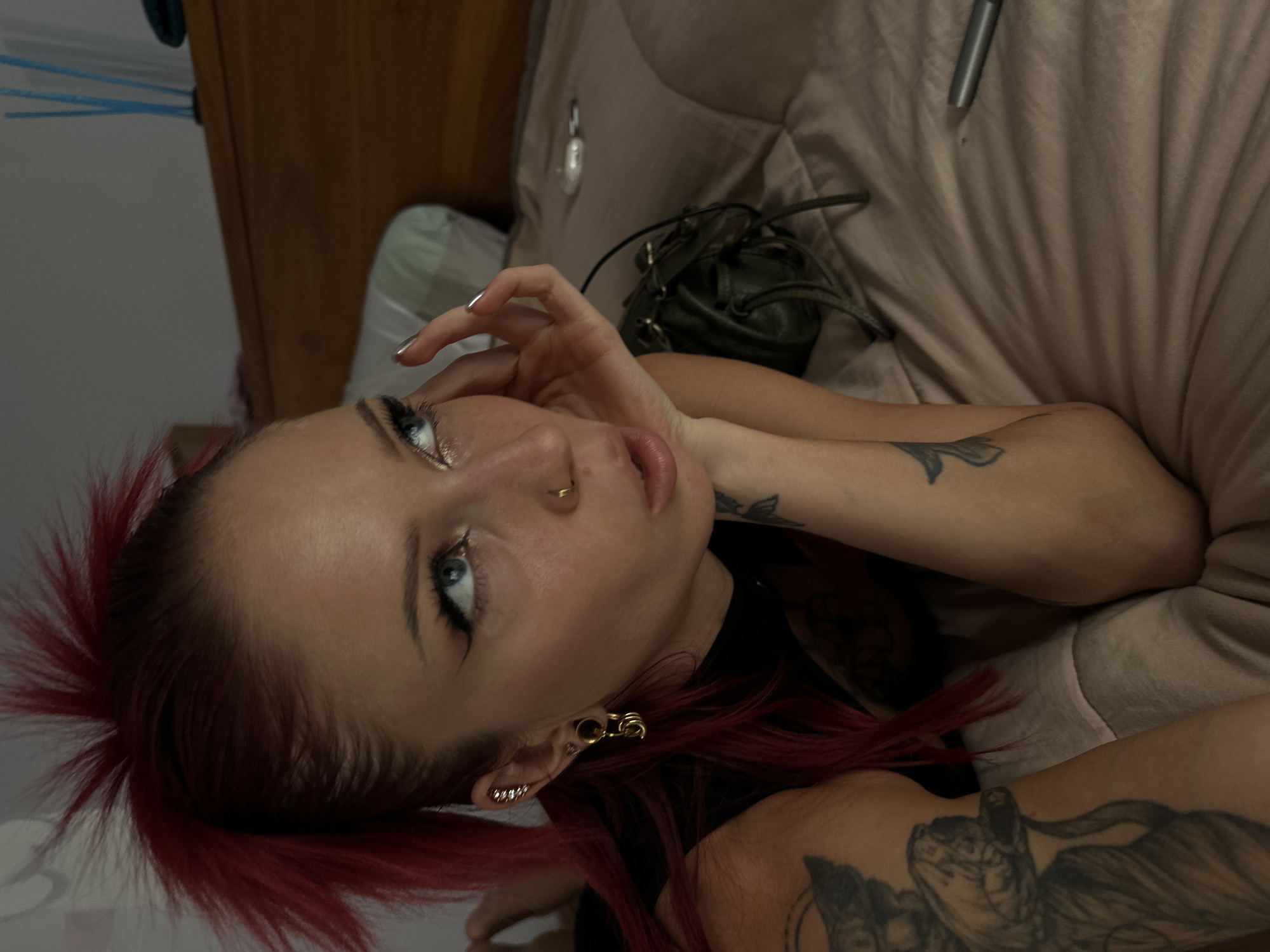 A woman lies on a beige bedspread, her head tilted toward the camera. She has fair skin, blue eyes, and red spiky hair. She has multiple piercings in her ear, including a gold-colored earring shaped like a safety pin. There is a nose ring in her left nostril. The woman's left hand is near her face, with silver nail polish. Her arms have tattoos. A small, dark green leather bag sits on the bedspread near her head. A silver-colored pen also rests on the bedspread. The location is implied to be in Dalung, Indonesia.