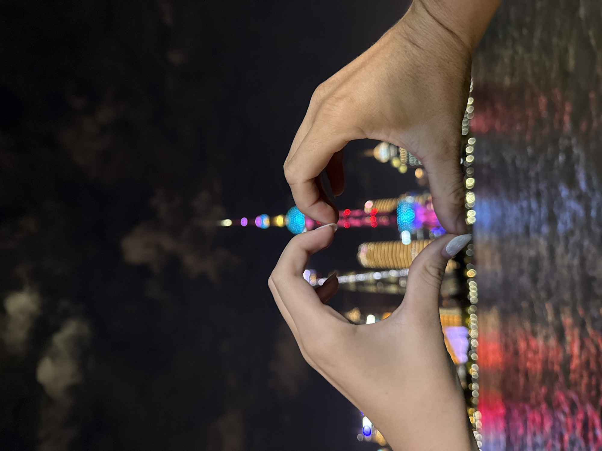 In this nighttime scene, two hands, one with light skin and a white-speckled fingernail, the other with darker skin, are brought together to form a heart shape. They are positioned in the foreground, with their thumbs touching and index fingers extended, creating the iconic symbol of love.
Behind the hands, the iconic Shanghai skyline is illuminated. The Oriental Pearl Tower, a prominent landmark in the Lujiazui area of the Pudong district, stands tall and is adorned with vibrant, colorful lights. Other skyscrapers in the background are also lit up, creating a glittering cityscape. The reflections of these lights shimmer on the dark water of the Huangpu River, which separates Pudong from Puxi. The water surface is textured with ripples, catching and distorting the bright reflections. The sky above is dark and cloudy, with some faint illumination suggesting the presence of distant lights or the moon obscured by clouds. The overall atmosphere is romantic and celebratory, capturing a moment of connection against the backdrop of a vibrant urban night. The location, with its distinctive skyline and river, is consistent with the Shanghai Bund or the Lujiazui financial district in the Pudong New Area, which is across the Huangpu River from Hongkou District, but the view is often associated with Shanghai's iconic waterfront.