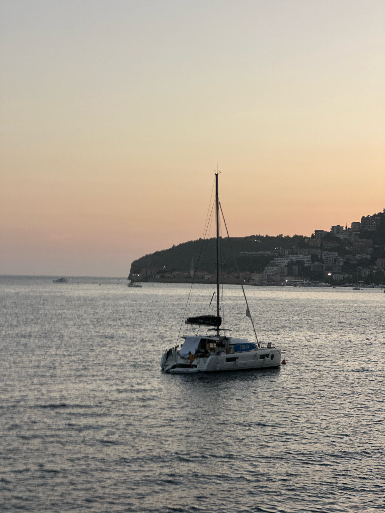 The image depicts a serene seascape at dusk, with a catamaran sailboat anchored in the foreground. The sailboat is white with dark sails and appears to be docked. In the background, the city of Budva, Montenegro, is visible along the coastline, with buildings nestled on a hillside. The sky is a soft gradient of orange and yellow, indicating the setting sun. The water is calm, with gentle ripples. The overall scene conveys a sense of peace and tranquility.