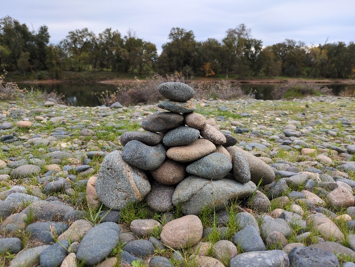 A stack of rounded rocks forms a small cairn on a riverbank. Various shades of grey and brown stones are piled together, surrounded by other scattered rocks and sparse patches of green grass. The background features a calm river, reflecting the sky, with a line of trees along the opposite bank. The sky is overcast, casting a soft light on the scene.