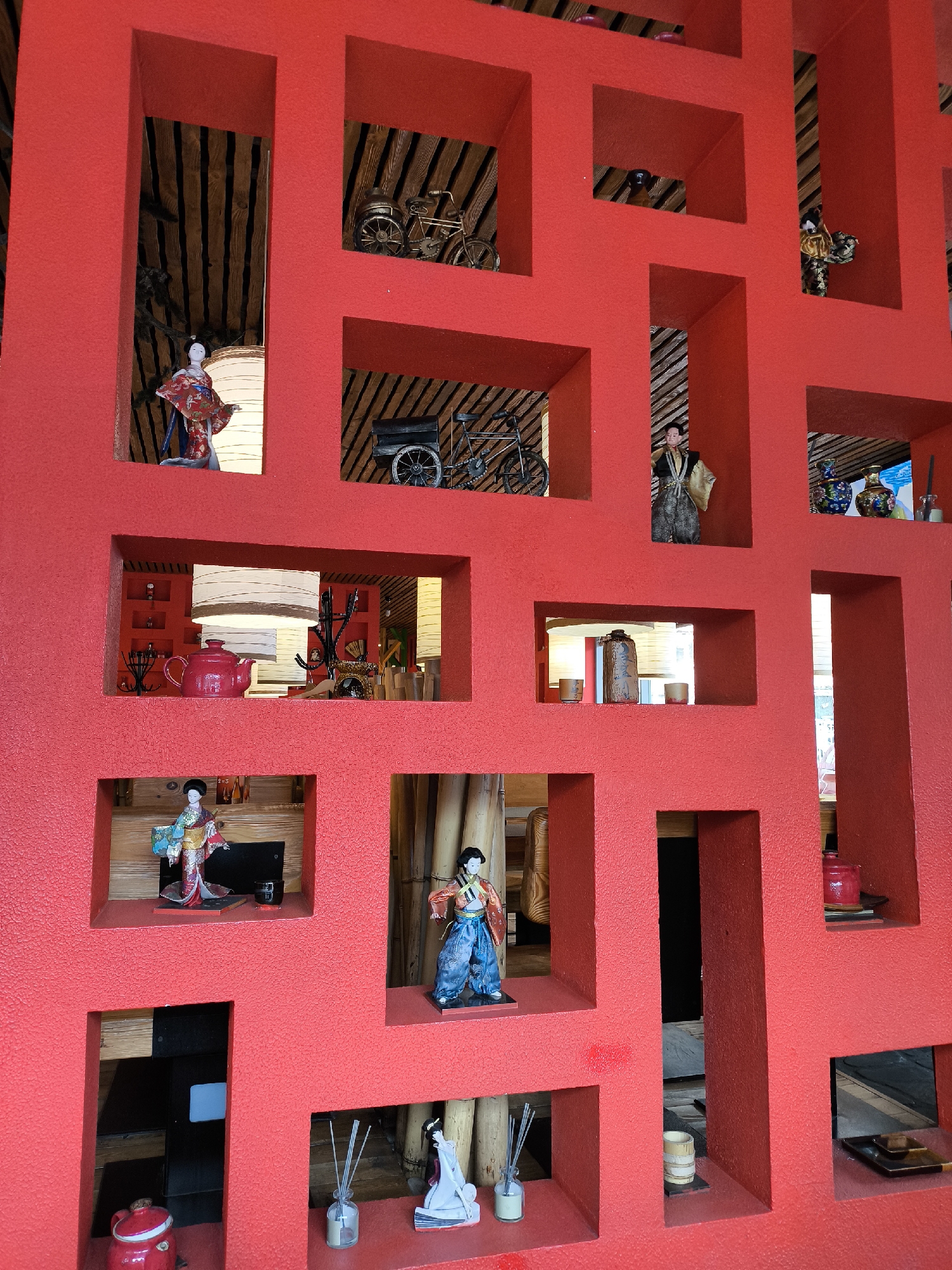 The image depicts a vibrant red, geometrically-structured display wall filled with various decorative items, likely within a restaurant or shop in Moscow, Russia. The wall is composed of numerous square and rectangular openings, each showcasing different objects.
These objects include:
* Several Japanese dolls in traditional attire.
* Miniature bicycles.
* Small decorative pots and teapots, predominantly red.
* A bottle of incense sticks.
* Other assorted decorative items such as small figurines and vessels.
The background visible through some of the openings suggests a textured ceiling with wooden beams, consistent with a rustic or themed interior. The red wall itself has a textured surface. The lighting appears to be a mixture of natural and artificial, with some sections illuminated brighter than others. The overall impression is one of carefully curated and organized display.