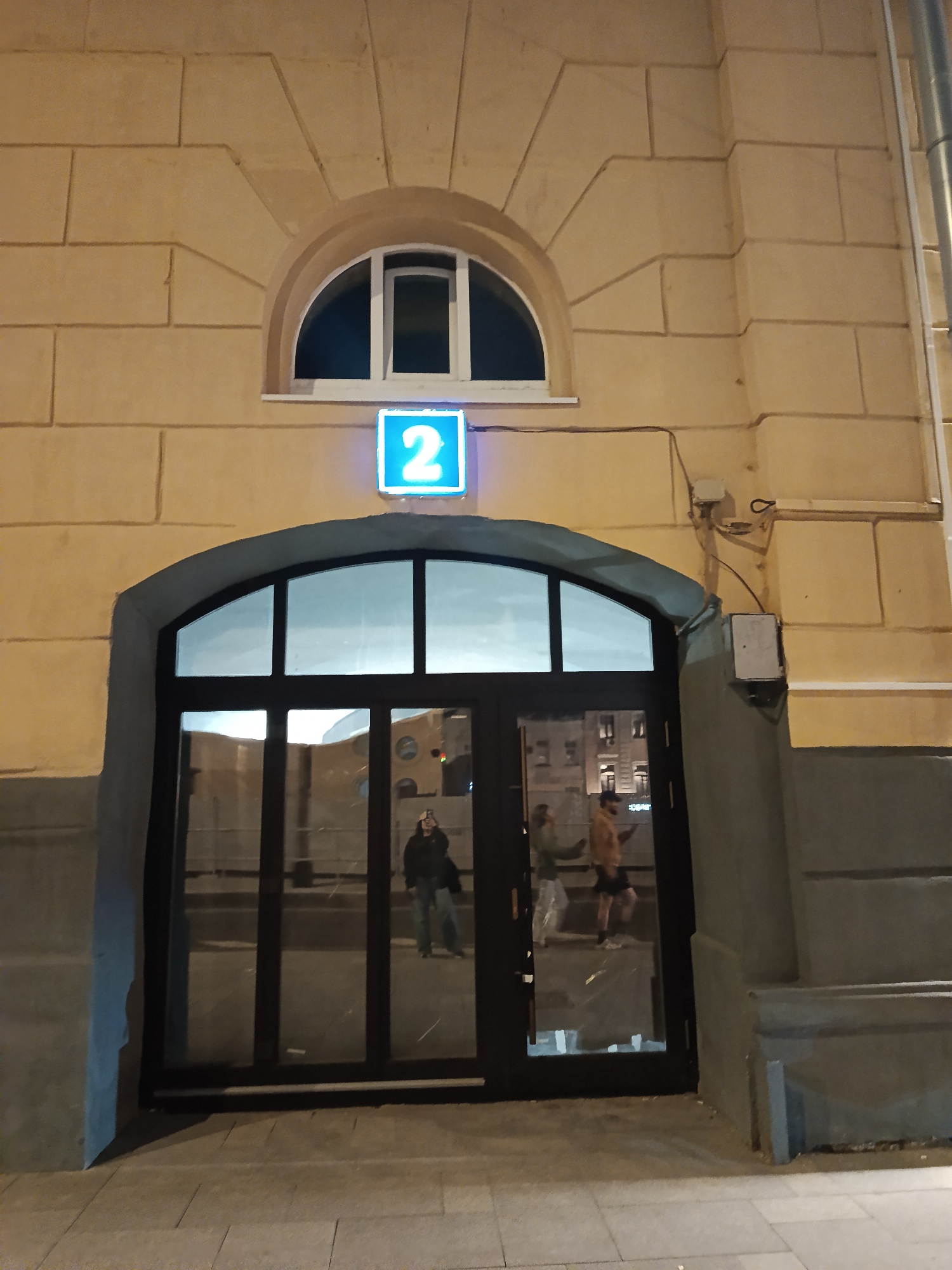 The photograph shows the exterior of a building, presumably at night, in an unidentified city. The architecture is notable for its arched doorway and an arched window above it. A blue, illuminated square with the number "2" is positioned between the window and the door. The building's facade is constructed from light-colored stonework. The arched doorway is flanked by darker, grey-toned stone columns. Inside the doorway, reflections reveal people and buildings. Two individuals are visible through the doorway's reflection, one on the left wearing casual clothes and one on the right wearing shorts and a jacket. The ground in front of the building is paved with light-colored rectangular stones.