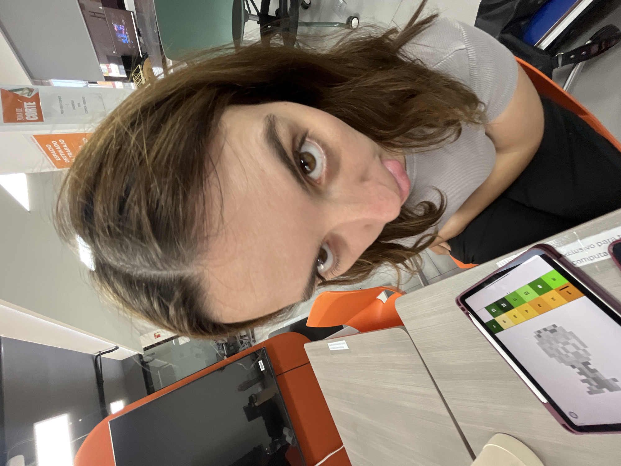 Here is a description of the image:
A tilted, close-up selfie depicts a young woman sticking her tongue out at the camera. She has brown hair with some gray strands and brown eyes. She is wearing a light gray shirt and seated in an orange chair. The background shows an office setting with cubicles and various screens. A tablet showing a digital image is on the desk in front of her. The phrases "ESPACIO DESTINADO TRABAJO", "ZONA DE CORTE" and "PROHIBIDO COMER E INGRESAR CON COMIDA Y BEBIDAS" are visible on the walls and panels, which suggests the photo was taken in San Andrés Cholula, Mexico.