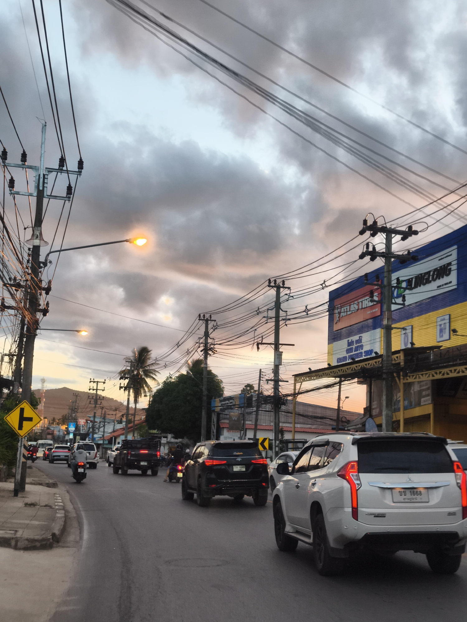 The image depicts a street scene in Baan Bo Phut, Thailand, during twilight. The sky is filled with dramatic clouds, with hints of orange and pink from the setting sun. Overhead, a dense network of electrical wires and utility poles dominates the upper portion of the frame, obscuring the sky in many areas.
On the right side of the street, a building with a large blue and yellow sign featuring the text "LINGLONG" and "ATLAS TIRES" is visible. Below this sign, a smaller sign reads "GRAND PREMIER SHOP".
The street itself is paved and appears to be a main thoroughfare with moderate traffic. Several vehicles are present, including cars, trucks, and motorcycles. In the foreground, a white SUV is prominently featured, its taillights illuminated. Behind it, a black SUV and other vehicles are also on the road. A motorcyclist is seen in the middle ground.
To the left of the street, a sidewalk with a yellow diamond-shaped traffic sign indicating a fork in the road is visible. Palm trees and other greenery are scattered throughout the scene, adding to the tropical environment. The overall impression is of a busy street in a coastal town as evening approaches.