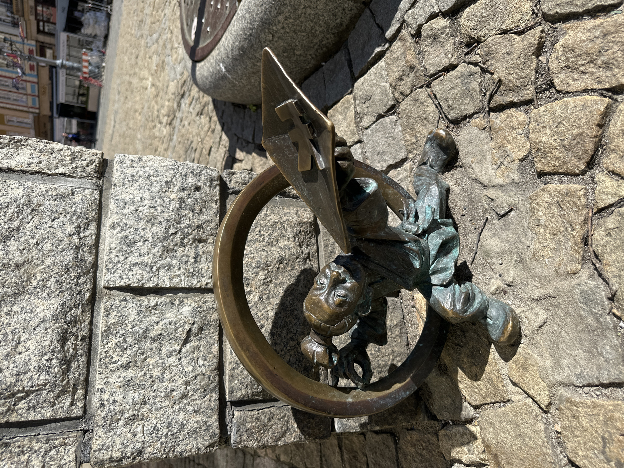 The image shows a bronze sculpture of a dwarf, part of the Wrocław Dwarves collection in Wrocław, Poland. The dwarf is depicted hanging upside down from a bronze ring, holding a shield with a cross on it. The sculpture is mounted on a stone wall. The lighting suggests a sunny day. In the background, there's a blurry urban scene with buildings and streets.