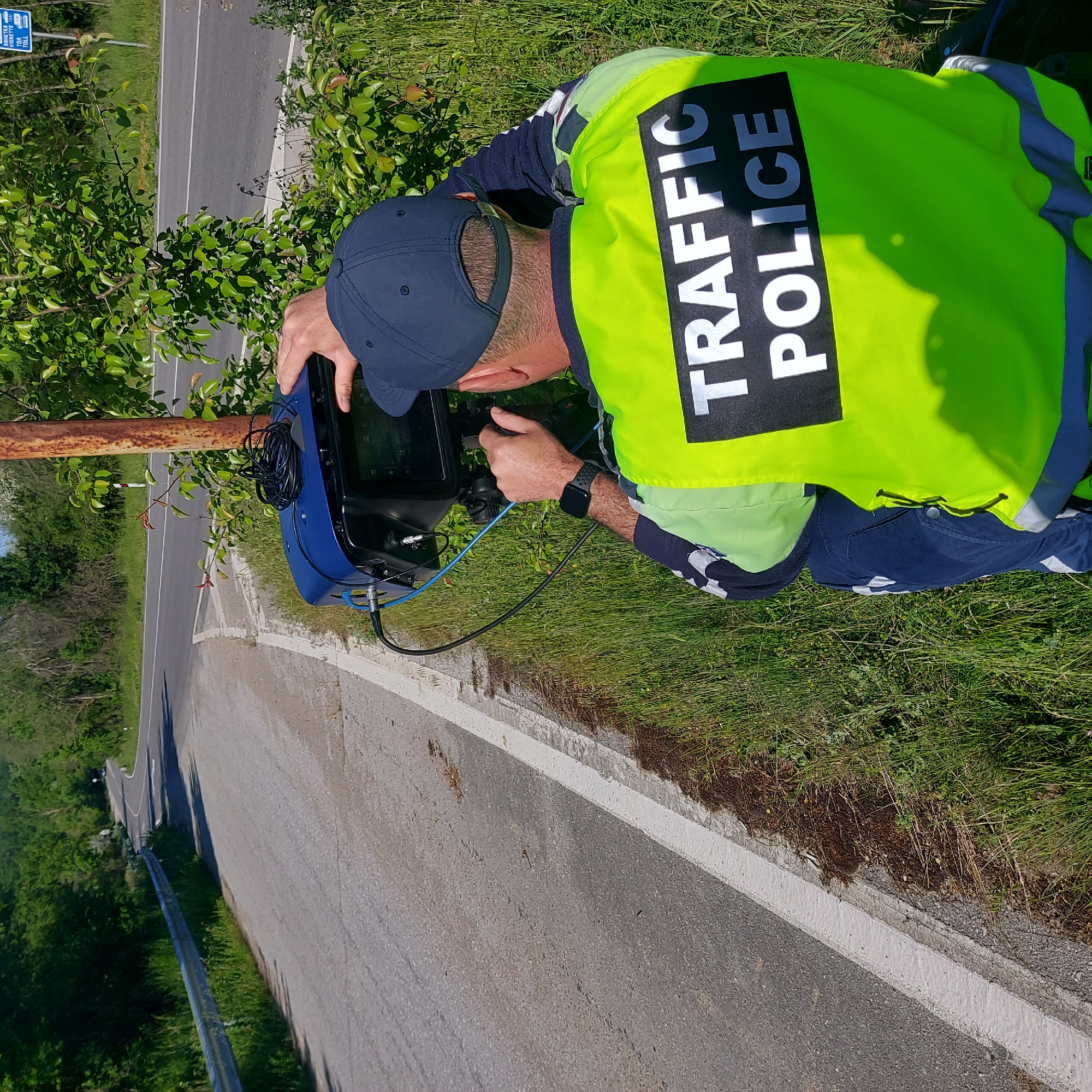 A traffic police officer, identified by the "TRAFFIC POLICE" inscription on their bright yellow vest, is operating a blue radar speed detection device positioned along a road. The location is indicated by a sign in the background, showing this is Balkantsi, Bulgaria. The officer is wearing a navy blue baseball cap, long-sleeved shirt, and pants. A black smartwatch is visible on their left wrist. The speed detection device is connected by cables, and the officer appears to be looking at the screen. The road is adjacent to a grassy area and some trees. A rusty metal guardrail is visible along the roadside.