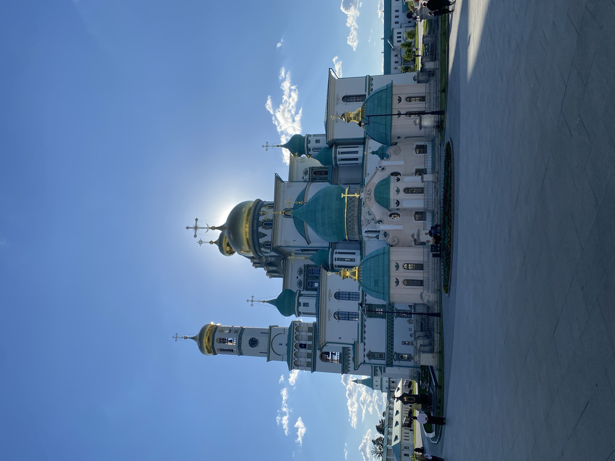 The image shows the New Jerusalem Monastery in Istra, Russia. The view is from a low angle, looking up at the complex. The sky is blue with some scattered white clouds. The sun is shining brightly behind the golden domes of the monastery, creating a halo effect. The architecture is a mix of white walls, green domes, and gold accents. The structures include a bell tower and several other buildings with detailed facades. In the foreground is a paved area. Some people are visible near the buildings on the right side of the image.