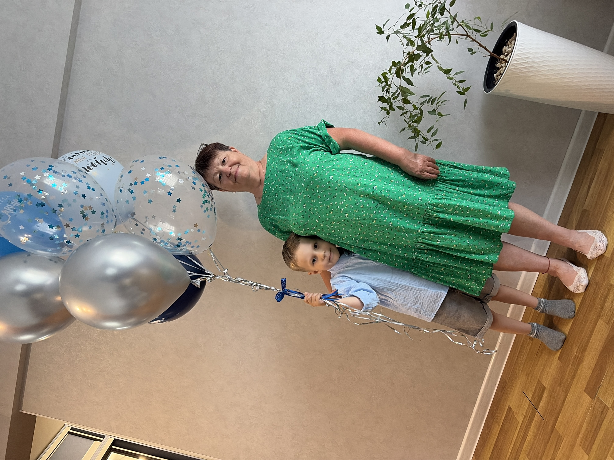 In this medium shot, a woman wearing a green floral dress and a young boy in a light blue collared shirt are the subjects, posed outdoors in what appears to be a bright and cheerful setting. They are surrounded by a cluster of balloons, primarily in shades of silver, blue, and white. Some of the balloons are clear and filled with blue confetti stars, while others are opaque silver and navy blue. The woman is looking towards the camera with a gentle smile, while the boy, peeking out from behind her, also gazes at the camera with a curious expression. His hand is visible, holding onto the strings of the balloons, which are adorned with blue ribbons and silver streamers.
The background reveals a textured light grey wall and a polished wooden floor. To the right, a white textured pot with a green leafy plant adds a touch of nature to the scene. The lighting suggests it is daytime, with no indication of specific weather conditions. The overall atmosphere conveyed is one of joy and celebration, possibly a birthday or a special occasion. The image has a tilted perspective, adding a dynamic quality to the composition. No visible text is present in the image.