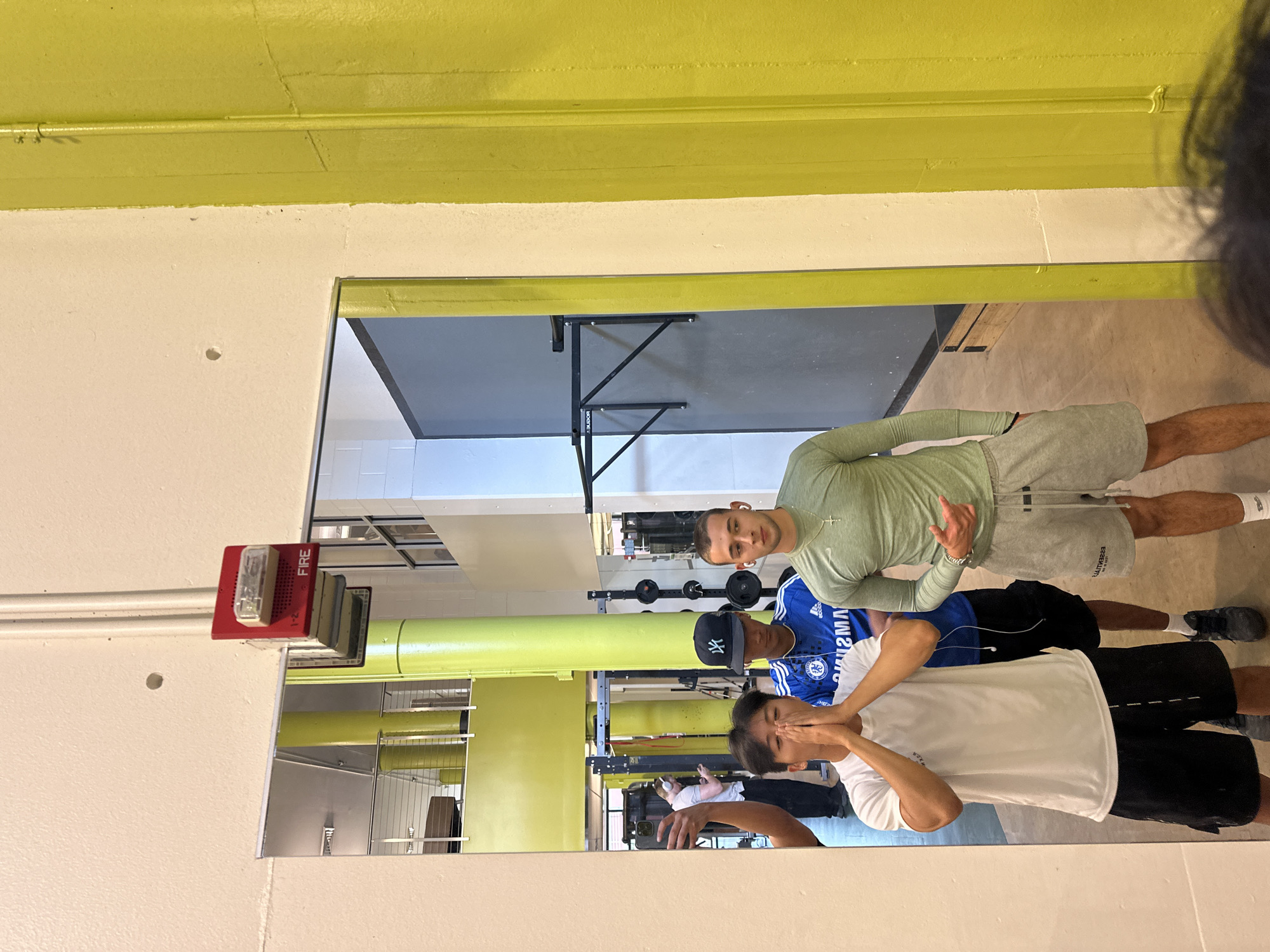 This image, taken in Tampa, United States of America, captures a mirror reflection of three young men posing in what appears to be a gym or fitness facility. The primary view is of the reflection, with a partial glimpse of the photographer's head in the top right corner.
In the reflection, one young man in a light green long-sleeve athletic shirt and grey shorts stands prominently, looking directly at the camera. He wears a silver cross necklace and a watch, and a hand gesture is visible. His shorts show the partially visible text "ESSENTIALS." To his left, slightly behind, is another young man wearing a blue football (soccer) jersey with "SAMSUNG" and the Adidas logo visible, black shorts, and a dark baseball cap with the "NY" logo of the New York Yankees. He is looking downwards. The third visible person in the reflection is the photographer, wearing a white t-shirt and black shorts, covering his face with both hands while holding a smartphone to capture the image.
The background reflected in the mirror shows typical gym equipment, including weight racks with barbells and plates. The walls in the facility are painted a bright lime green on the upper section and white below. A red fire alarm unit, labeled "FIRE," with a clear strobe light, is mounted on the wall to the left of the reflection. A wall-mounted foldable table, with the word "BACKER" partially visible on its leg, is also reflected. The floor appears to be a light-colored, possibly concrete, surface. The scene depicts a casual moment among friends in a gym environment.