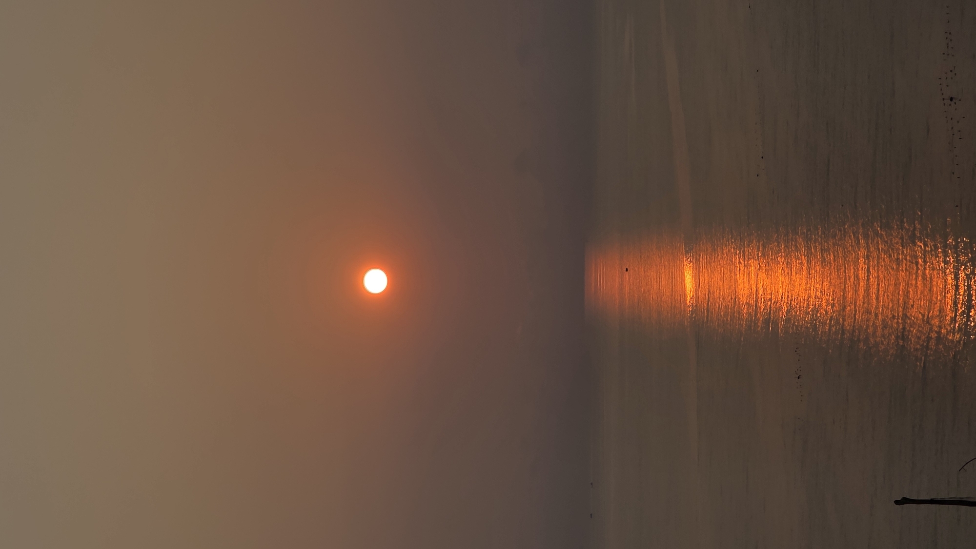 The image shows a sunset over water, likely in Bac Natural Heritage Area Trang, Vietnam. The sun is a bright orange orb in the left half of the image, obscured by a hazy atmosphere. The water surface reflects the sun's light in a bright, elongated band extending towards the bottom right. The overall color palette is dominated by muted orange and gray tones, suggesting a polluted or smoky atmosphere.