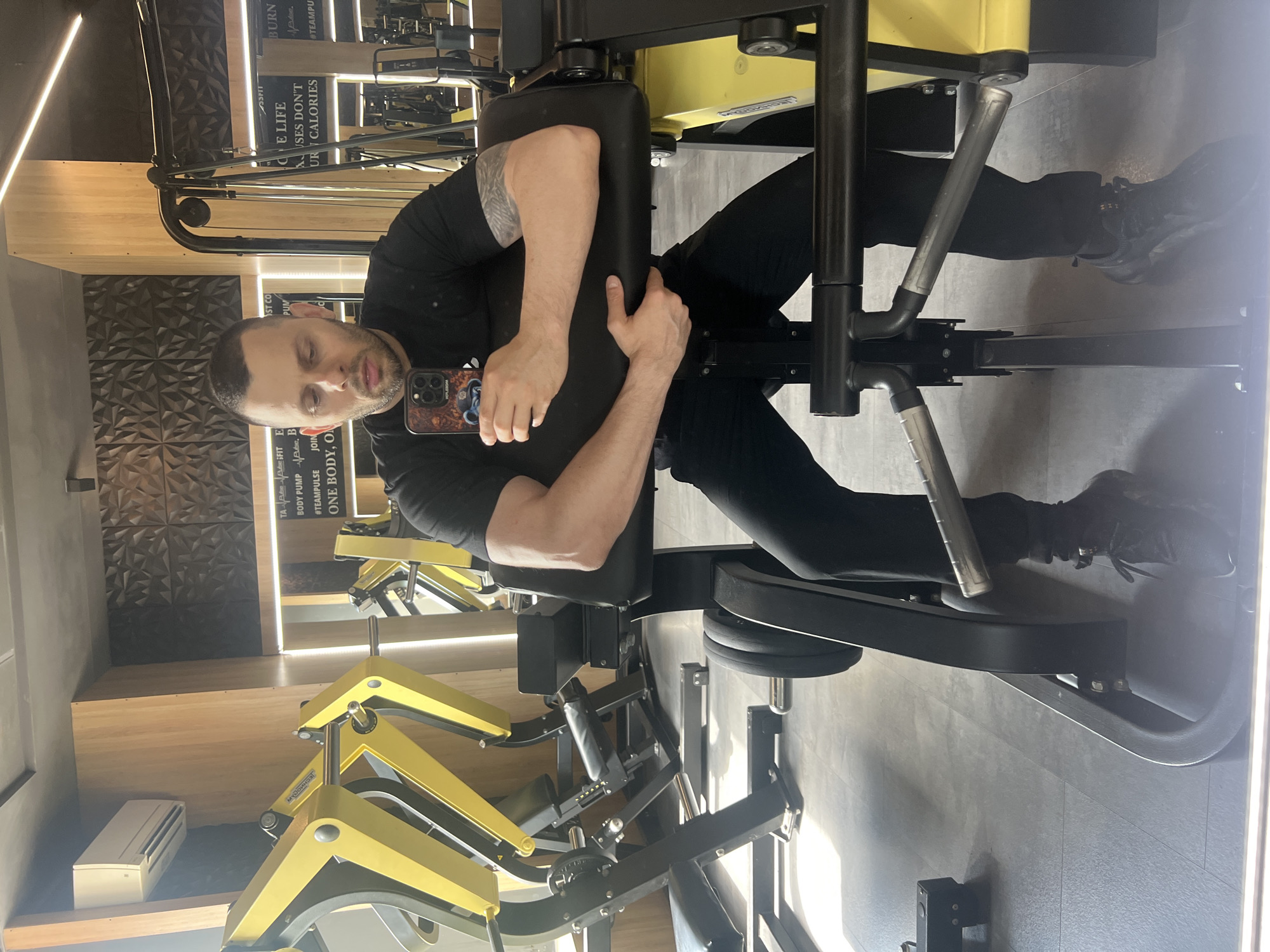 A man is sitting in a leg extension machine at a gym. He is wearing a black shirt and black pants. He is holding his phone in his hand, and appears to be taking a selfie. The gym appears to be in Burgas, Bulgaria, based on the Cyrillic text on the wall and the "Myodom" logo on the machine. The wall has motivational phrases written on it, such as "BURN," "ONE BODY, ON." and "LIFE." The gym has light wood and grey tile flooring, and the gym equipment is black and yellow.