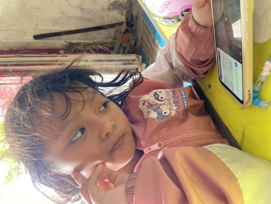 A young child with dark hair looks pensively to the side, touching their cheek. They wear a pink and white jacket with a "Kuromi Sweet Baby" design. A yellow phone is held nearby, displaying a screen with text. The background is cluttered, with glimpses of boxes and a metal bar. The image is rotated counter-clockwise, presenting an angled perspective. Beads of blue and white are attached to the phone.