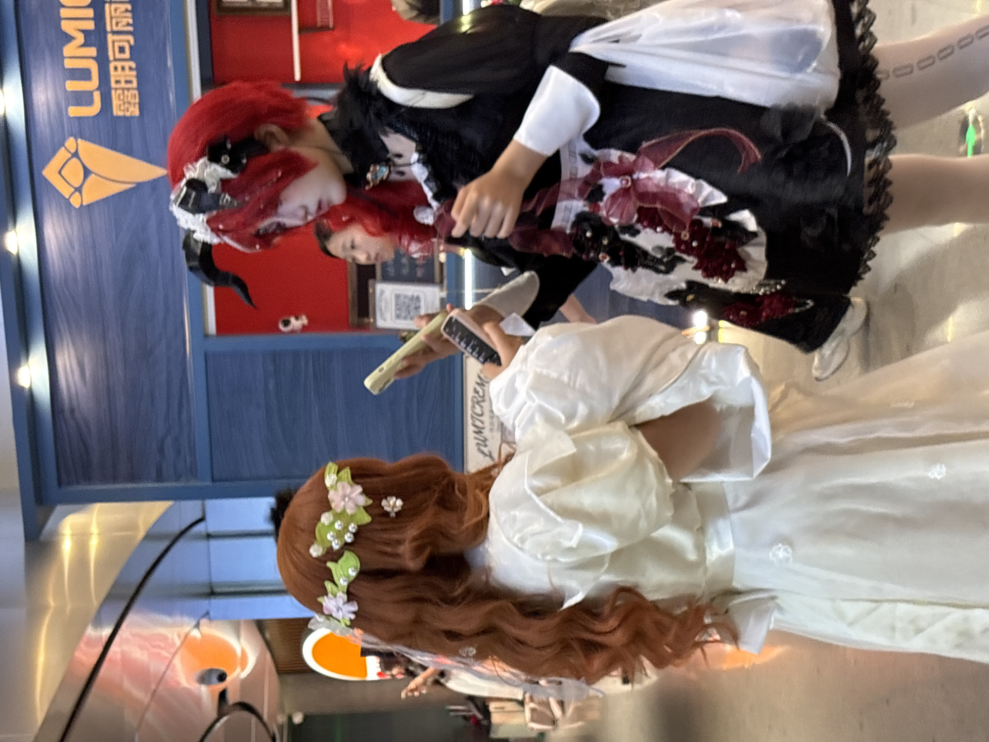 Two individuals are captured in the image. One, dressed in a black and white Lolita-style dress with red hair and what appears to be a black horn, is facing another person dressed in a white dress with red hair adorned with a floral headpiece. The scene seems to be taking place in an indoor area, possibly a mall or shopping center, suggested by the presence of store signs and the reflective flooring. The location appears to be Unknown, Unknown. They are holding a phone, taking a video of each other.