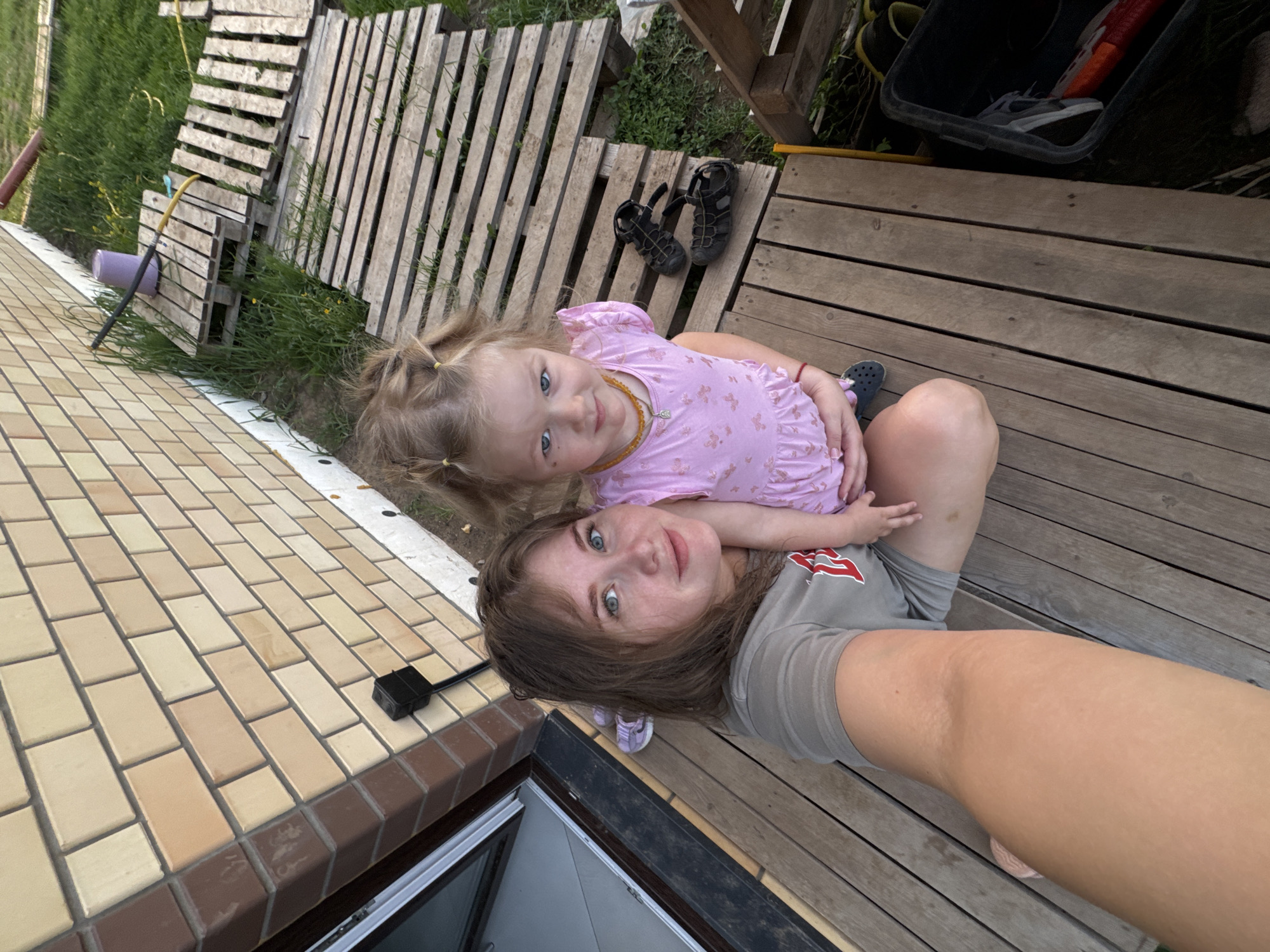 A high-angle shot shows a woman and a young girl outside a brick building and a wooden deck, potentially in the city of Ферма, Russia. The young girl, wearing a pink dress with butterfly patterns and her blonde hair in pigtails, lies on top of the woman, who is wearing a gray t-shirt. The woman is looking up at the camera. A pair of children's sandals lie on the deck, and a wheelbarrow is visible in the upper right corner. The building features tan bricks on the facade and a dark-framed window is visible. Adjacent to the deck is an area with wooden pallets laid out on the grass.