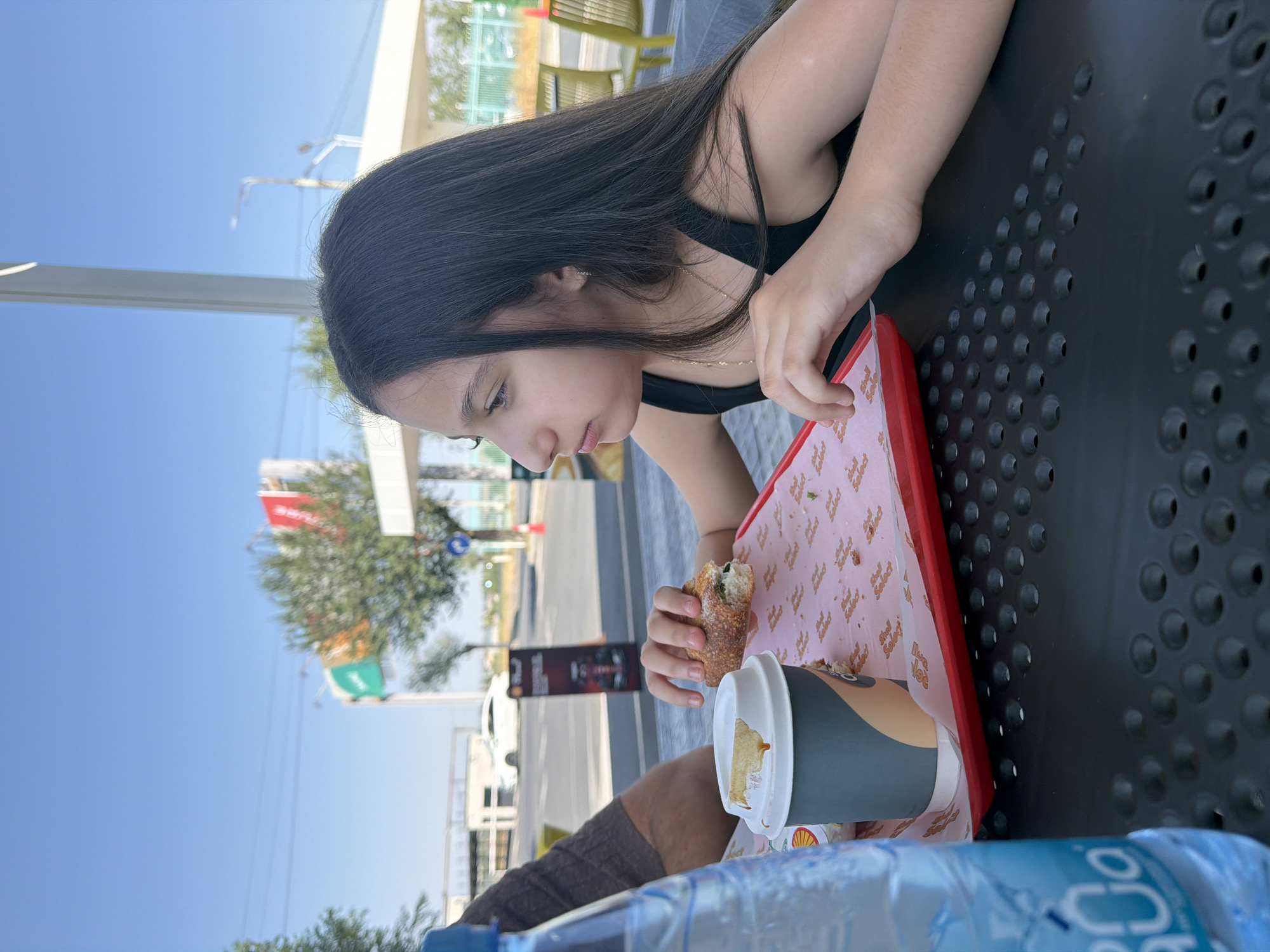 A young girl with long, dark hair sits at an outdoor table, eating a sandwich. She is wearing a black tank top and a gold necklace. In front of her is a red tray with the sandwich and a disposable coffee cup. To her left, a partially visible person's arm in a brown sweater is resting on the table. A clear plastic water bottle with blue lettering is also on the table, to the left of the frame. The table is dark gray with a pattern of circular indentations. In the background, there are trees, buildings, a road with cars, and a blue sky with overhead wires. The scene appears to be a public outdoor seating area, possibly a rest stop or food establishment, located in Parakar, Armenia.