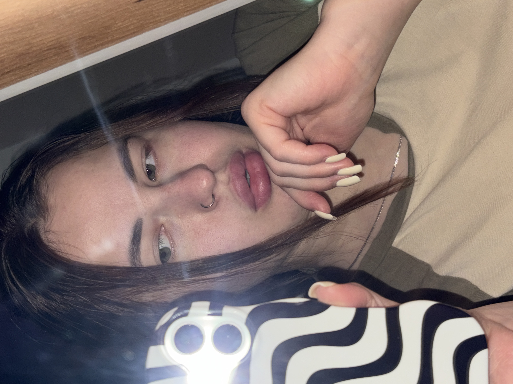 A young woman is shown in a selfie. She is lying down and holding a phone with a black-and-white striped case. The woman has brown hair and fair skin. She has a nose ring and is wearing a necklace. She is wearing a tan-colored shirt. The background includes a wooden shelf and a green garment. The location of the image is unknown.