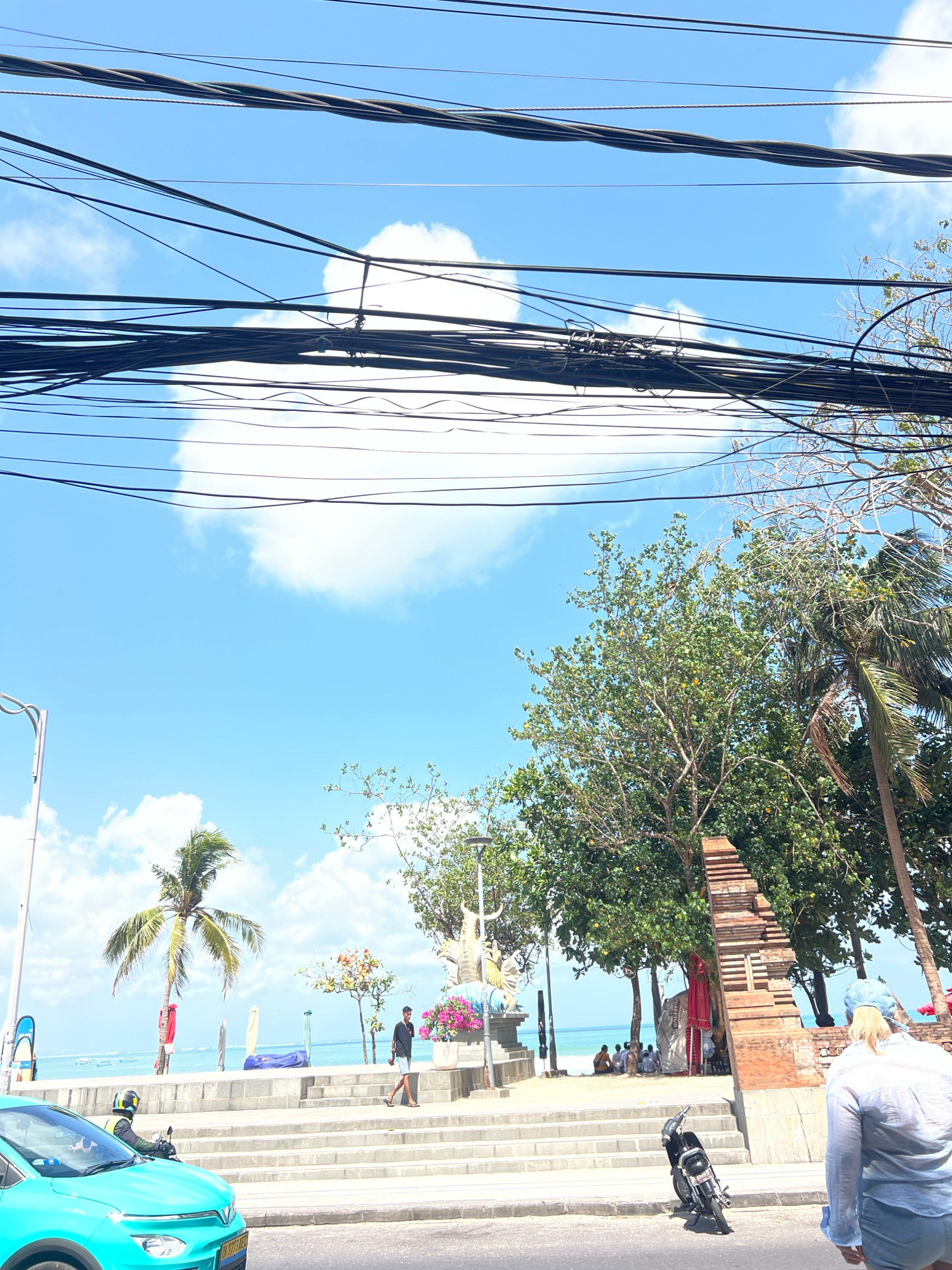 In Kuta, Indonesia, a bright, sunny day unfolds along a coastal road. Overhead, a dense network of black electrical wires crisscrosses the vivid blue sky, punctuated by fluffy white clouds. Below the wires, a palm tree stands tall near the beach, where several surfboards lean against a wall. A man in a black shirt and shorts walks down a set of concrete stairs towards the sand, where a group of people are gathered. To the right, a brick structure resembling a temple entrance stands amidst lush green trees. A turquoise car is visible on the left side of the frame, with a person on a motorcycle in the driver's seat. Further down the road, a motorcycle is parked on the pavement, and a person wearing a light blue shirt and denim shorts walks away from the camera. The scene captures a typical day in this popular Indonesian tourist destination.
