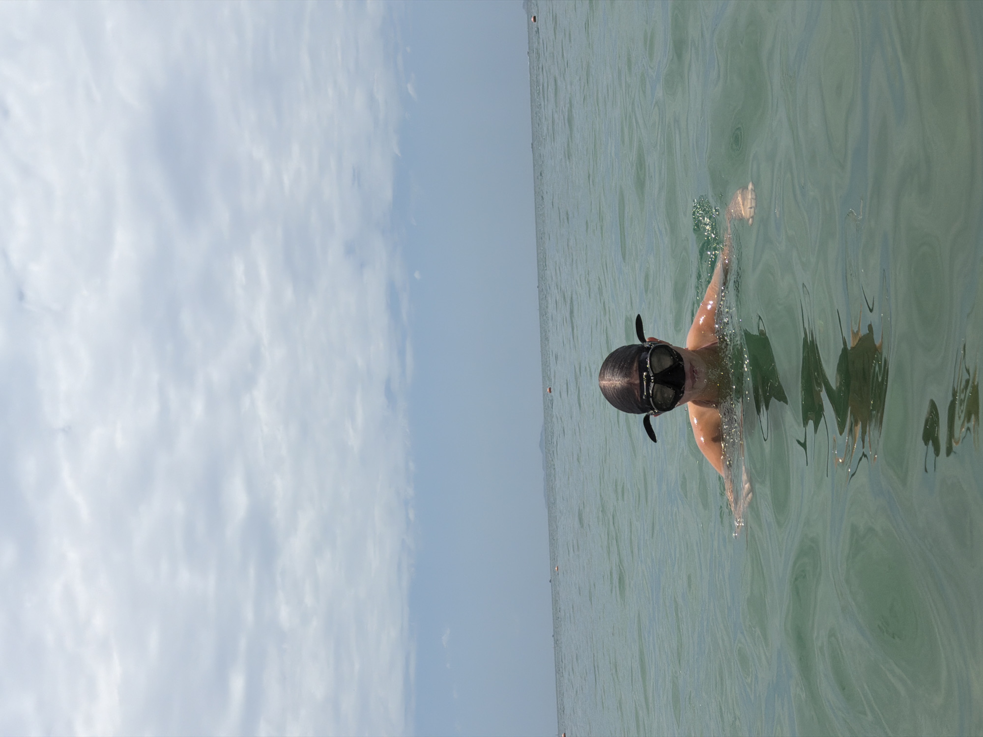 A person wearing a black diving mask and snorkel is partially submerged in the ocean in Nha Trang, Vietnam. The water is green and appears to be calm with slight ripples. The sky above is overcast with a light gray cloudy texture. In the distance, a hazy landmass is visible on the horizon, with several small orange buoys floating in the water. The person is facing forward, looking directly at the camera, with their head and shoulders above the water.