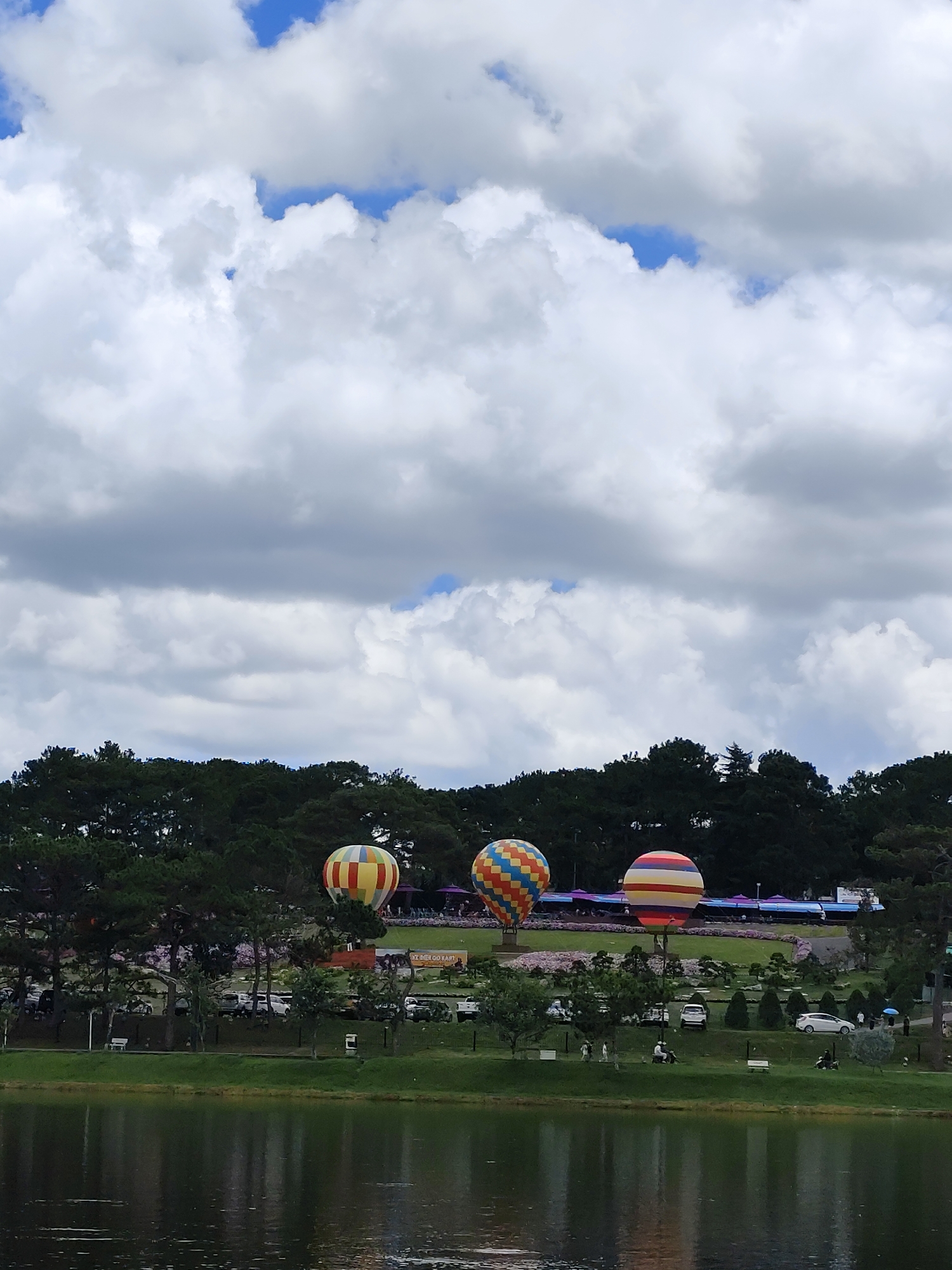 The image shows a scenic view of Xuan Huong Lake in Da Lat, Vietnam. In the foreground, a calm lake reflects the overcast sky and the surrounding greenery. Across the lake, a lush green landscape is dotted with trees and manicured gardens. Three colorful hot air balloons are prominently displayed, appearing to be part of an event or festival. The balloons are of varying sizes and patterns: one is yellow and red, another is a vibrant orange, yellow, and blue checkered design, and the third has horizontal stripes of red, yellow, and white. Behind the balloons, a line of trees forms a dense backdrop against the cloudy sky. The sky itself is filled with dramatic, cumulus clouds, with patches of blue sky visible between them. There are people present on the grass near the balloons, along with some vehicles parked in the background. The overall scene depicts a lively outdoor gathering set against a picturesque natural backdrop.