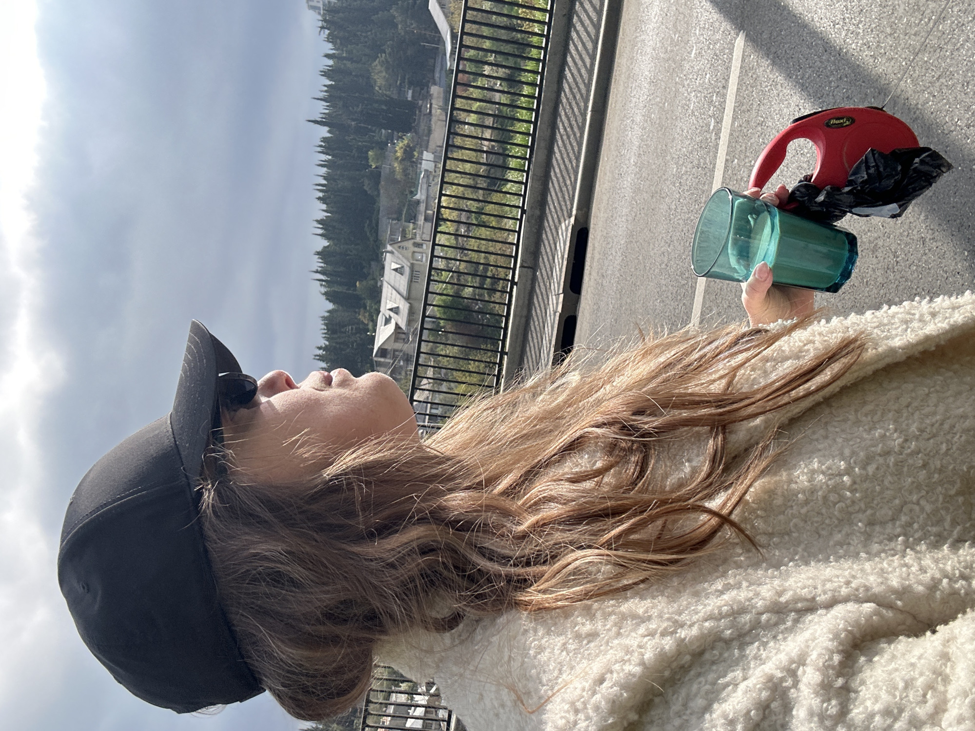 A woman with long brown hair wearing a black baseball cap and a white fleece jacket is holding a teal glass and a red dog leash attached to a black plastic bag. The background is a cityscape with trees, buildings, and a bridge railing. The location is likely Tbilisi, Georgia.