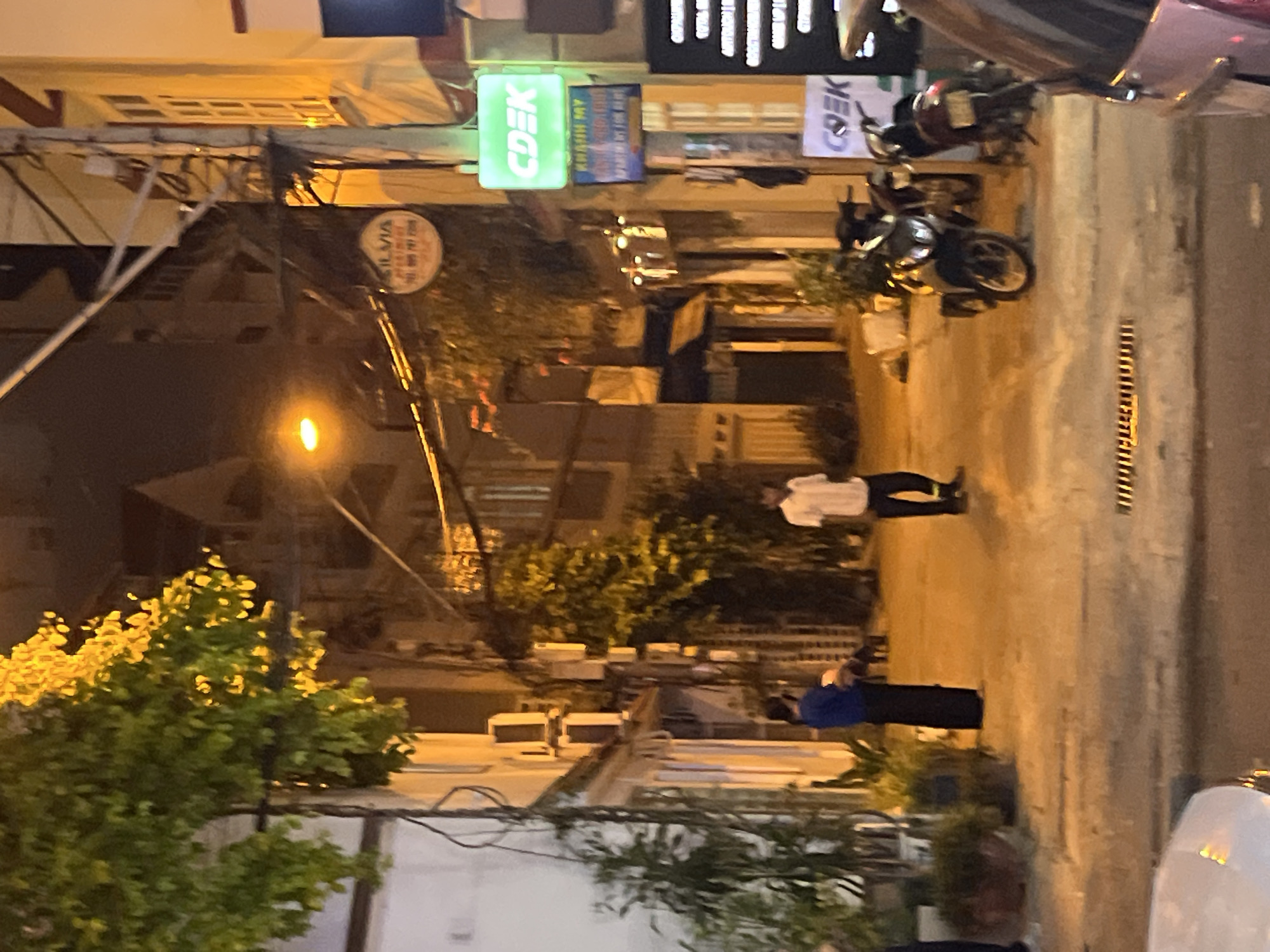 A nighttime street scene in Bac Nha Trang Ward, Vietnam, features a narrow, dimly lit alleyway with buildings on either side. Green foliage hangs from a tree in the upper left. Two people are visible on the walkway: one in a blue top and dark skirt, and another in a white short-sleeved shirt and dark pants. Several motorcycles are parked along the right side of the alley. Signs for "CDLK" and "KHANH MY APARTMENT FOR RENT" are illuminated. The overall lighting is orange and yellow, suggesting streetlights and building illumination.