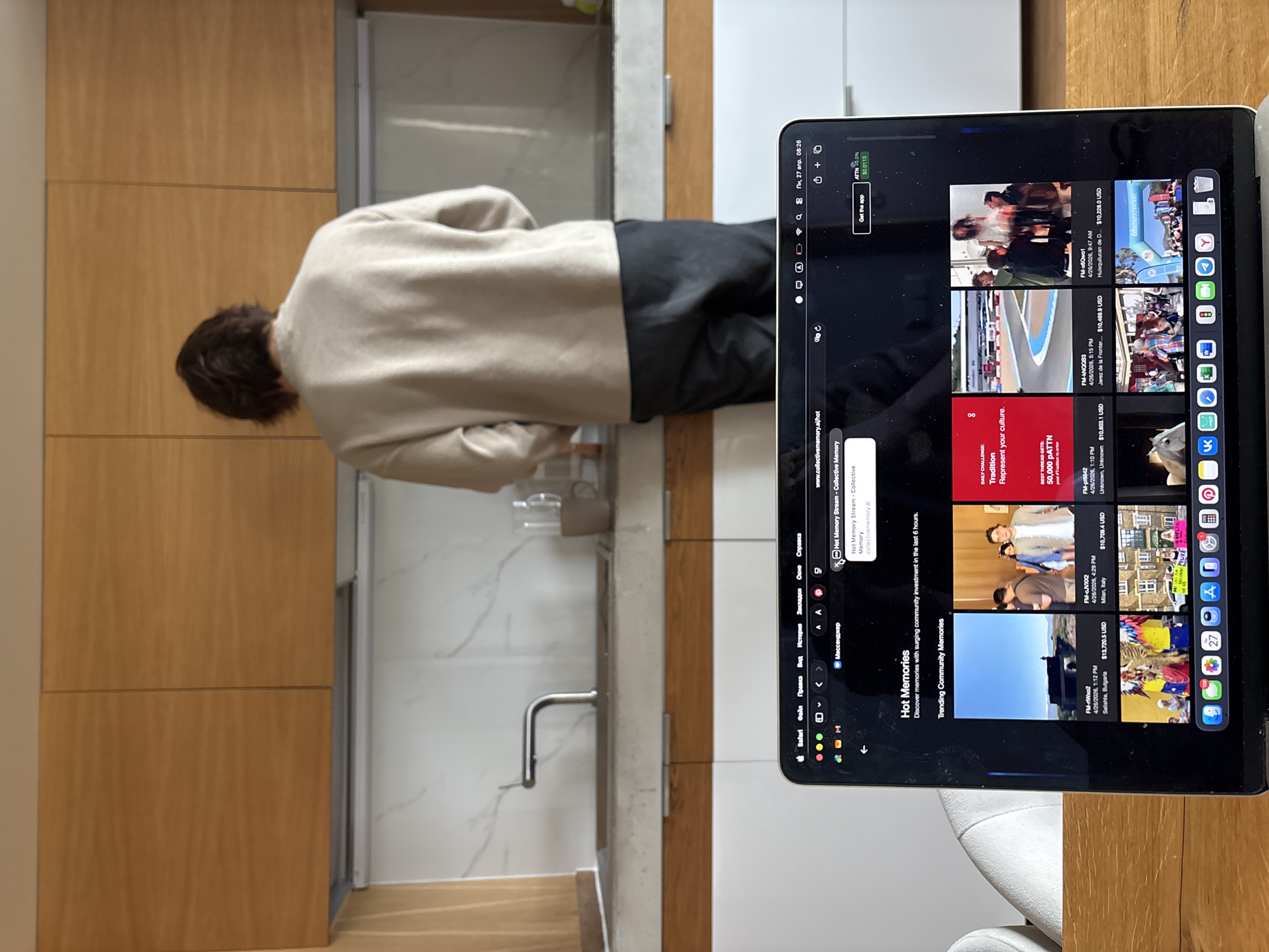 A person stands at a kitchen sink next to an iPad. The iPad displays the website "www.collectivememory.al/hot," showcasing various trending community memories. The kitchen has wooden cabinets and a stainless steel faucet. The location is not possible to determine from the information.