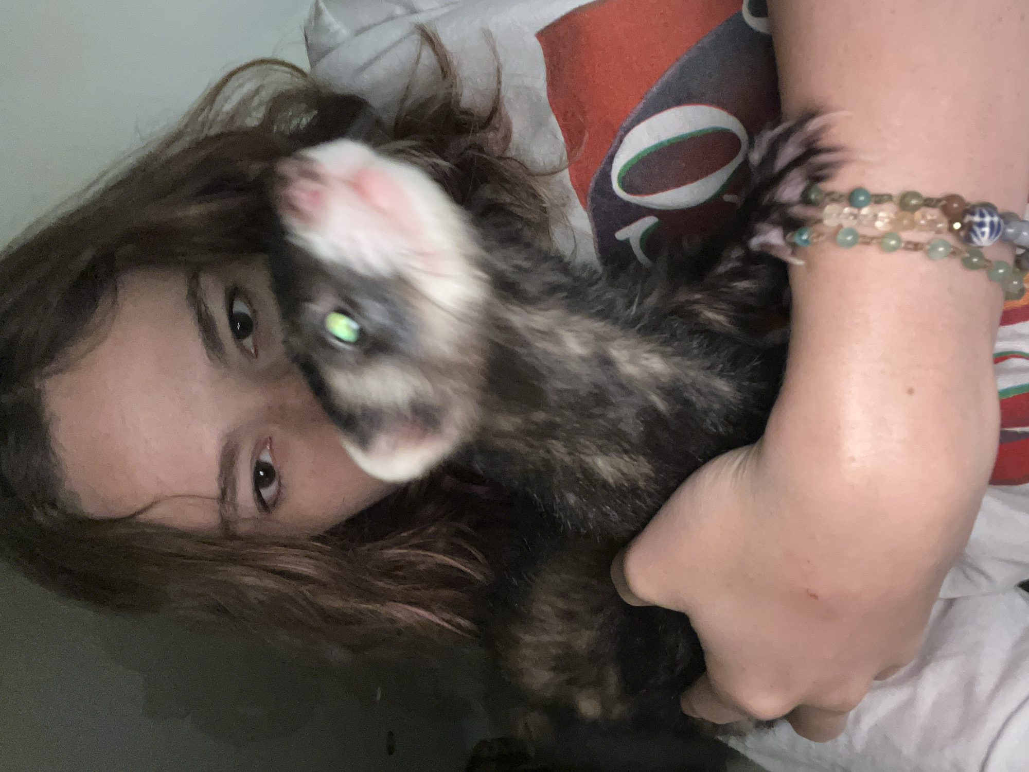 In San Bernardino Tlaxcalancingo, Mexico, a close-up, dimly lit indoor scene features a person holding a ferret. The individual, with brown, wavy hair, has their face partially obscured by the animal, revealing only their right eye. They are wearing a white t-shirt with a red and dark-colored graphic design visible on the right shoulder/chest area. The person's left hand and forearm are holding a dark-furred ferret, which exhibits a mottled pattern of black, brown, and tan. The ferret's head is slightly blurred, indicating motion, with a pink nose, white fur around the muzzle, and prominent green eye shine. Its light-colored front paws are visible. A multi-strand beaded bracelet, featuring various green, blue, brown, and clear beads, including one blue and white patterned bead, is worn on the person's left wrist. A small reddish mark is present on the back of the left hand. The background is dark and out of focus.