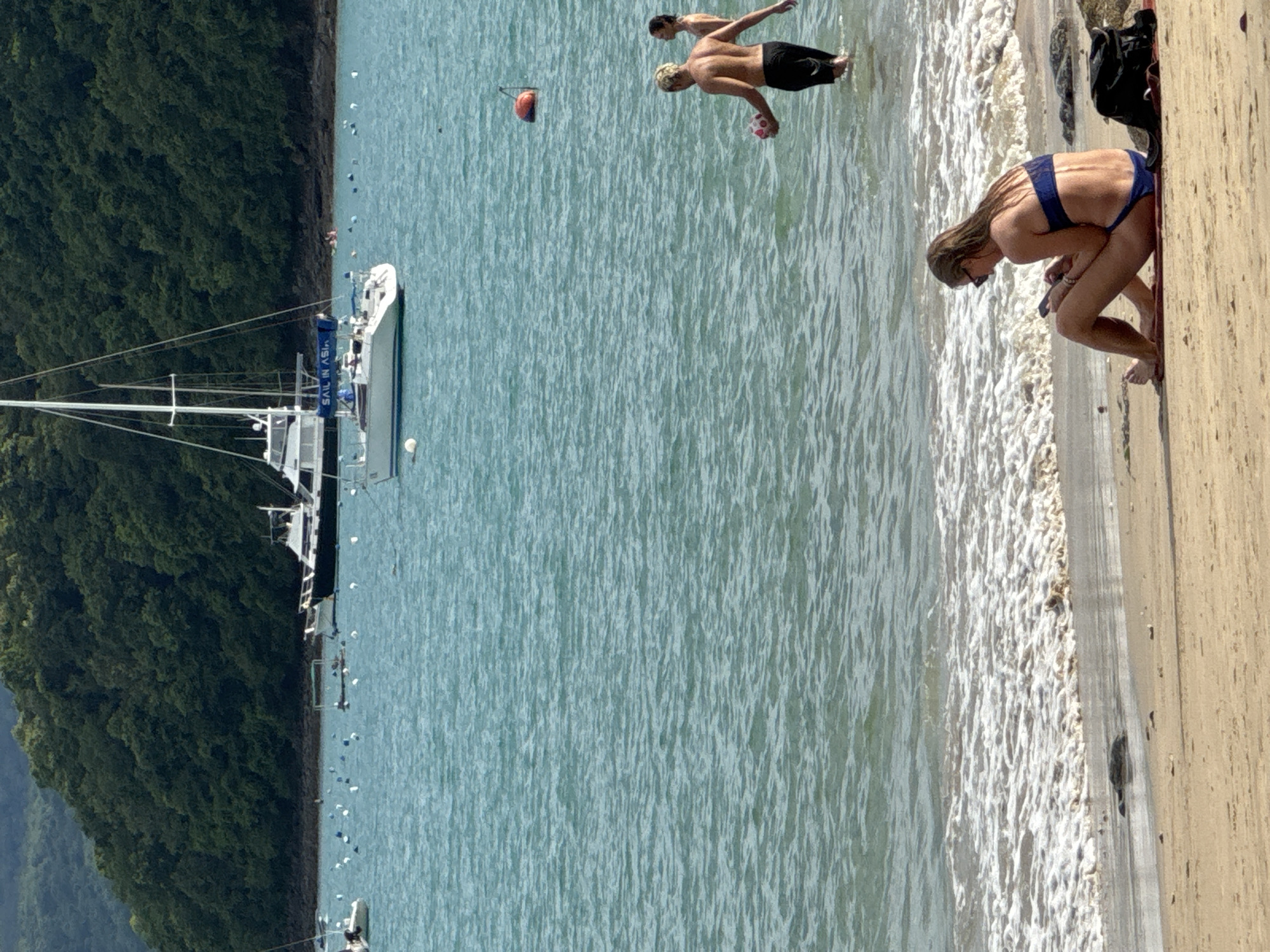 A sunlit, medium shot captures a tropical beach scene in Wichit, Thailand. In the foreground, a woman with long, blonde hair, wearing sunglasses and a blue bikini, sits on a red towel on the sand. She is looking down at her phone, which she holds in her hands. To her right, a black bag rests on the sand. The gentle waves of the turquoise ocean lap at the shore behind her.
In the middle ground, two individuals, likely a couple, stand in the shallow water. The person in front, with blonde hair and a muscular back, is wearing black swim trunks and holds a pink and white ball. The person behind them, partially obscured, appears to be wearing swimwear.
Further out in the water, several boats are anchored. A white sailboat with "SAIL IN ASIA" written on its blue sail is prominently featured. In the distance, a larger white yacht is visible, as well as other smaller boats and buoys. The background is dominated by a lush, green, tree-covered hillside, characteristic of the coastal landscape in Wichit. The water is clear and calm, reflecting the bright sky.