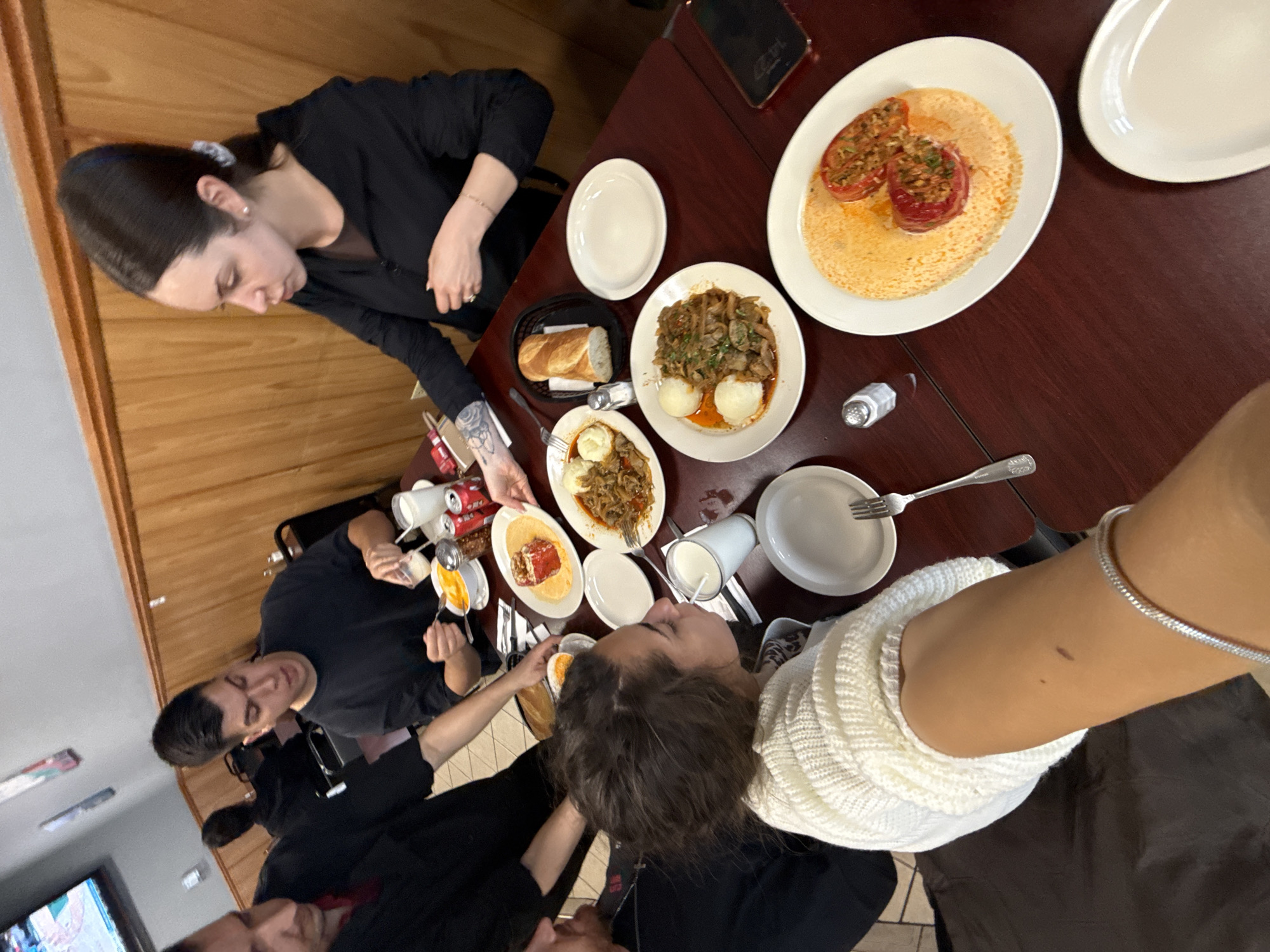 The image captures a dining scene at a restaurant in Des Plaines, Illinois. A group of people are seated around a dark wood-grained table laden with food. A woman is serving food to another person at the table. Dishes include stuffed tomatoes and a mushroom-based dish. Other items such as bread, beverages, and condiments are visible. The restaurant has wood paneled walls, and the atmosphere is casual.