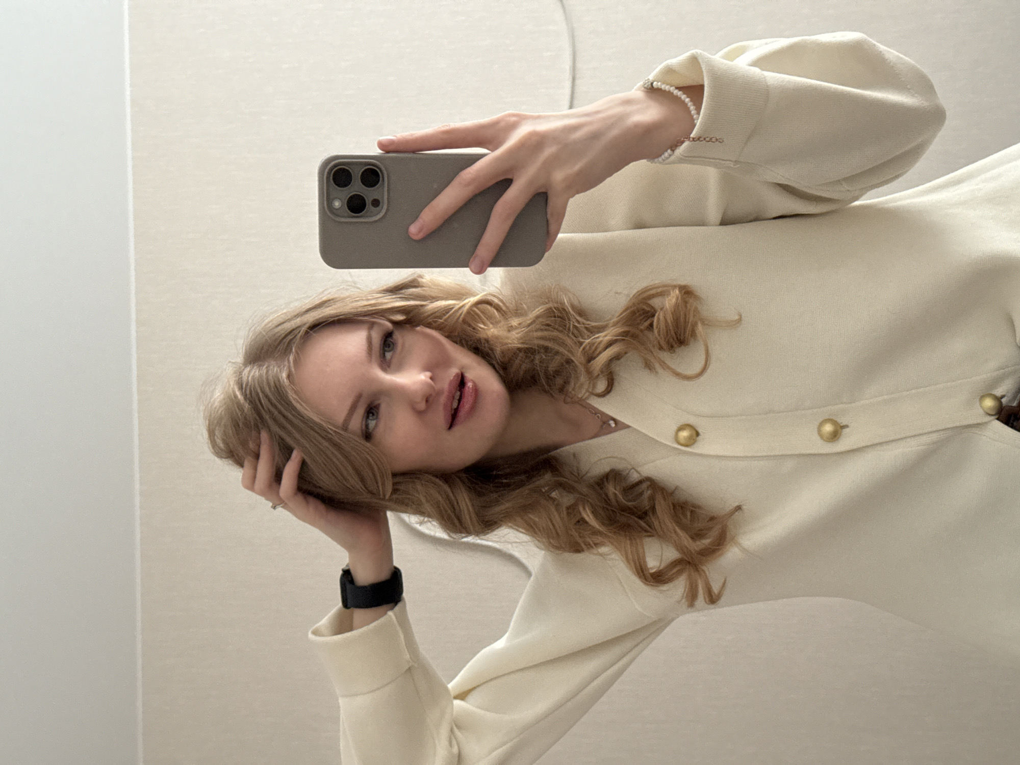 A woman is taking a selfie in a bathroom. The woman has light blonde hair and is wearing a cream-colored cardigan with gold buttons and a black smartwatch on her left wrist. She is holding a cell phone in her right hand, with a tan-colored case. The background consists of a neutral-colored wall, likely within a bathroom setting. There are no details confirming this is Perm, Russia.
