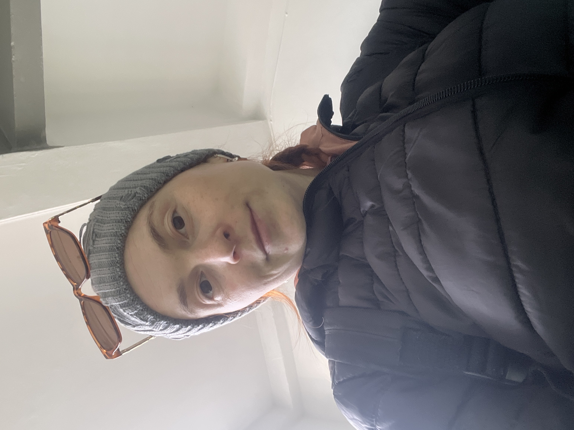 A low-angle selfie shot appears to have been taken in a likely indoor location in Chaikovsky, Russia. The subject is a person wearing a gray knit beanie with a pair of sunglasses perched on it. They are also wearing a puffy black jacket. The background is a bright, neutral color, possibly an interior wall. No specific details allow for identifying the subject beyond the general clothing and accessories. No further context can be inferred about interactions or actions.
